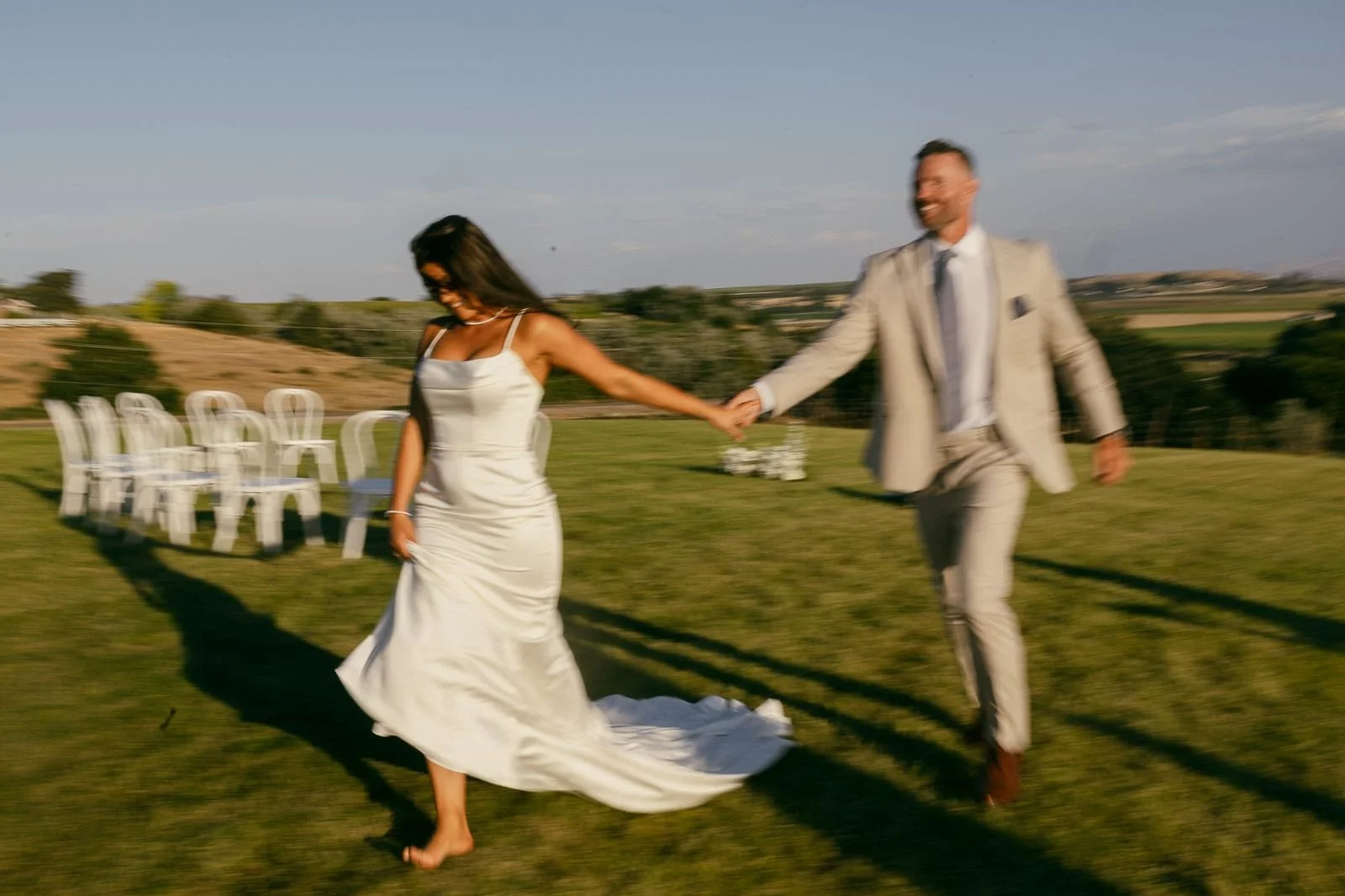  A bride in a white dress and a groom in a light suit hold hands and walk barefoot on grass, smiling, with empty white chairs and a scenic landscape in the background. The image is slightly blurred, conveying motion. 