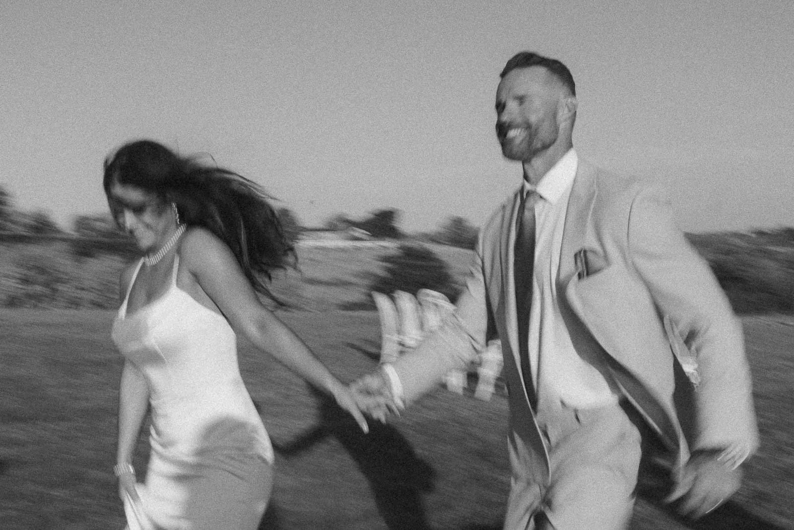 A black-and-white photo showing a smiling couple in formal attire holding hands and running outdoors, with blurred motion conveying excitement and joy.