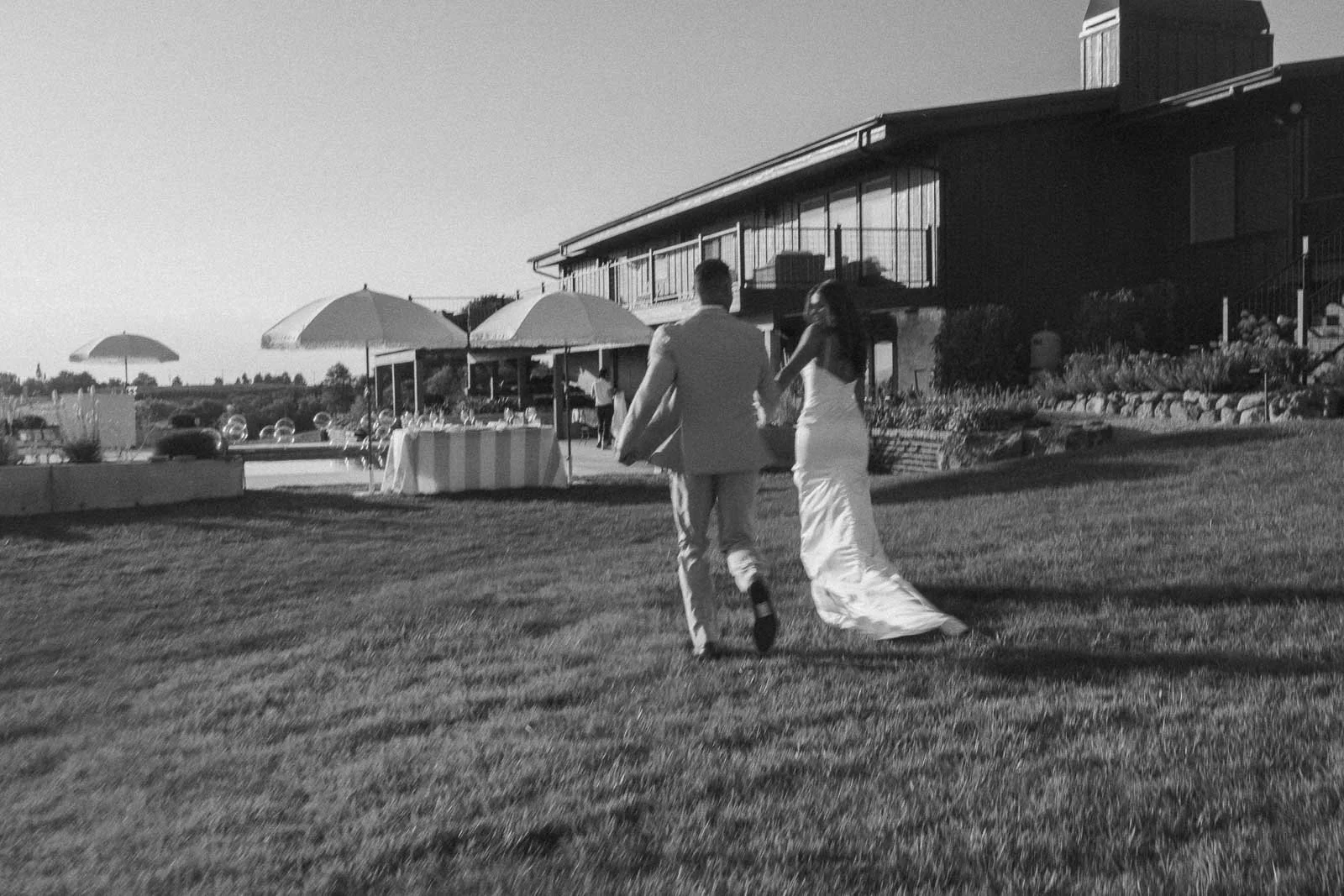  A bride and groom, seen from behind, hold hands and run across a grassy lawn toward an outdoor event area with umbrellas and a large building in the background. The image is in black and white. 
