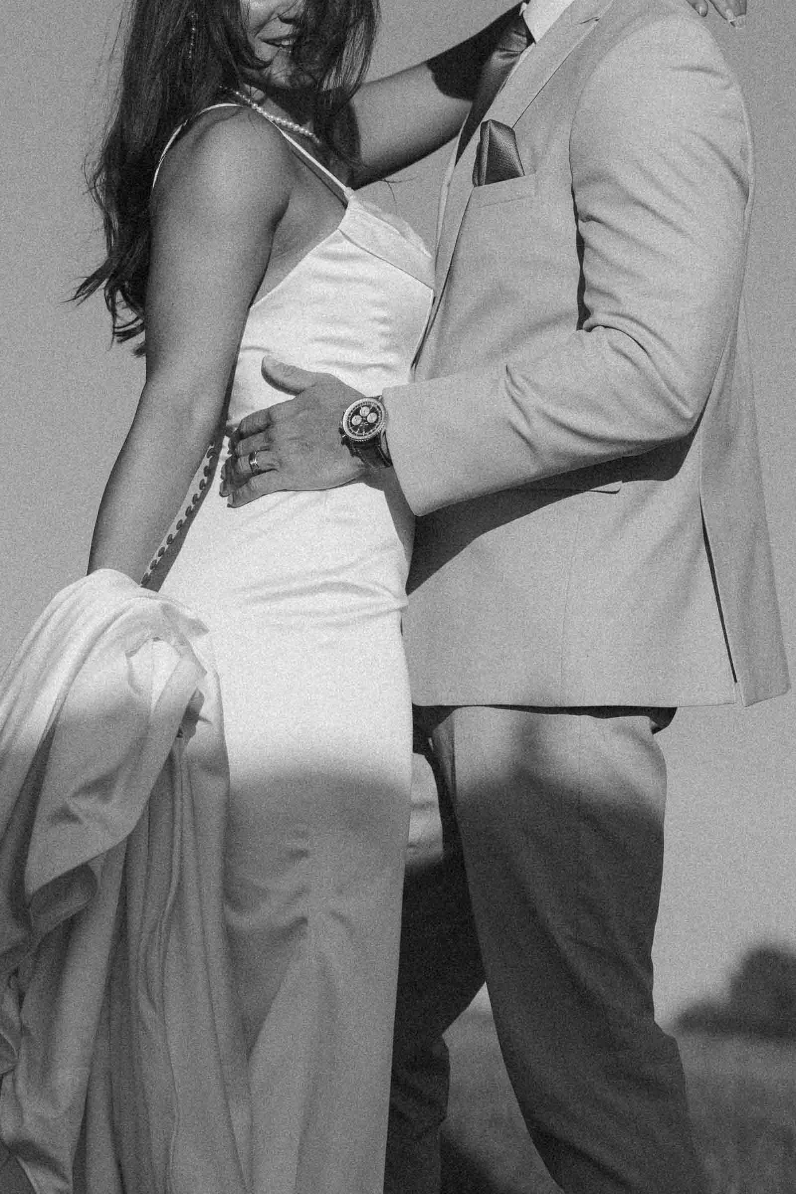  A black-and-white photo of a couple dressed formally, with the woman in a satin gown and the man in a light suit. They are standing close together outdoors, with the man's hand on the woman's waist. 