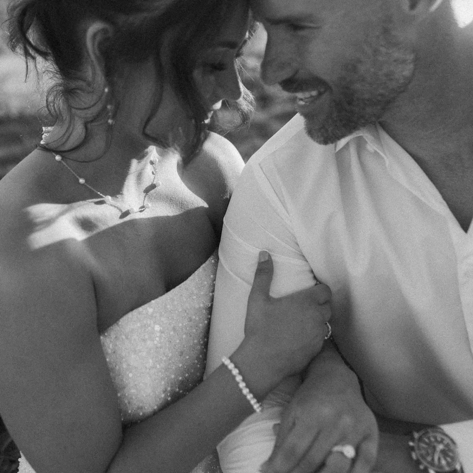 A black and white photo of a bride and groom smiling closely together. The bride is holding the groom’s arm, showing her engagement ring, and they both look happy and affectionate. 