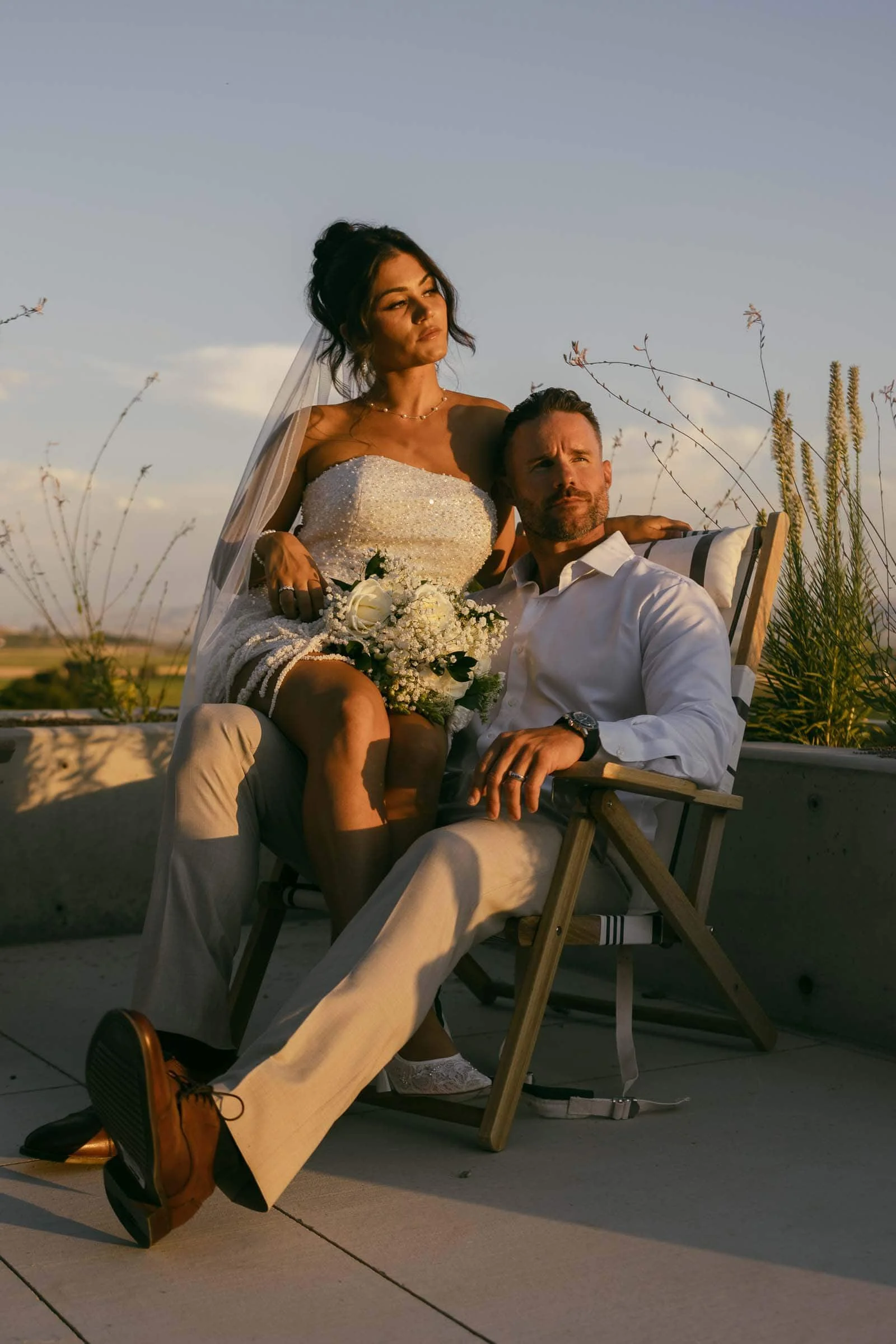  A bride in a white dress and veil sits on the lap of a groom in light-colored clothes, holding a bouquet, both looking off into the distance. They are outdoors with plants and a sunny sky in the background. 