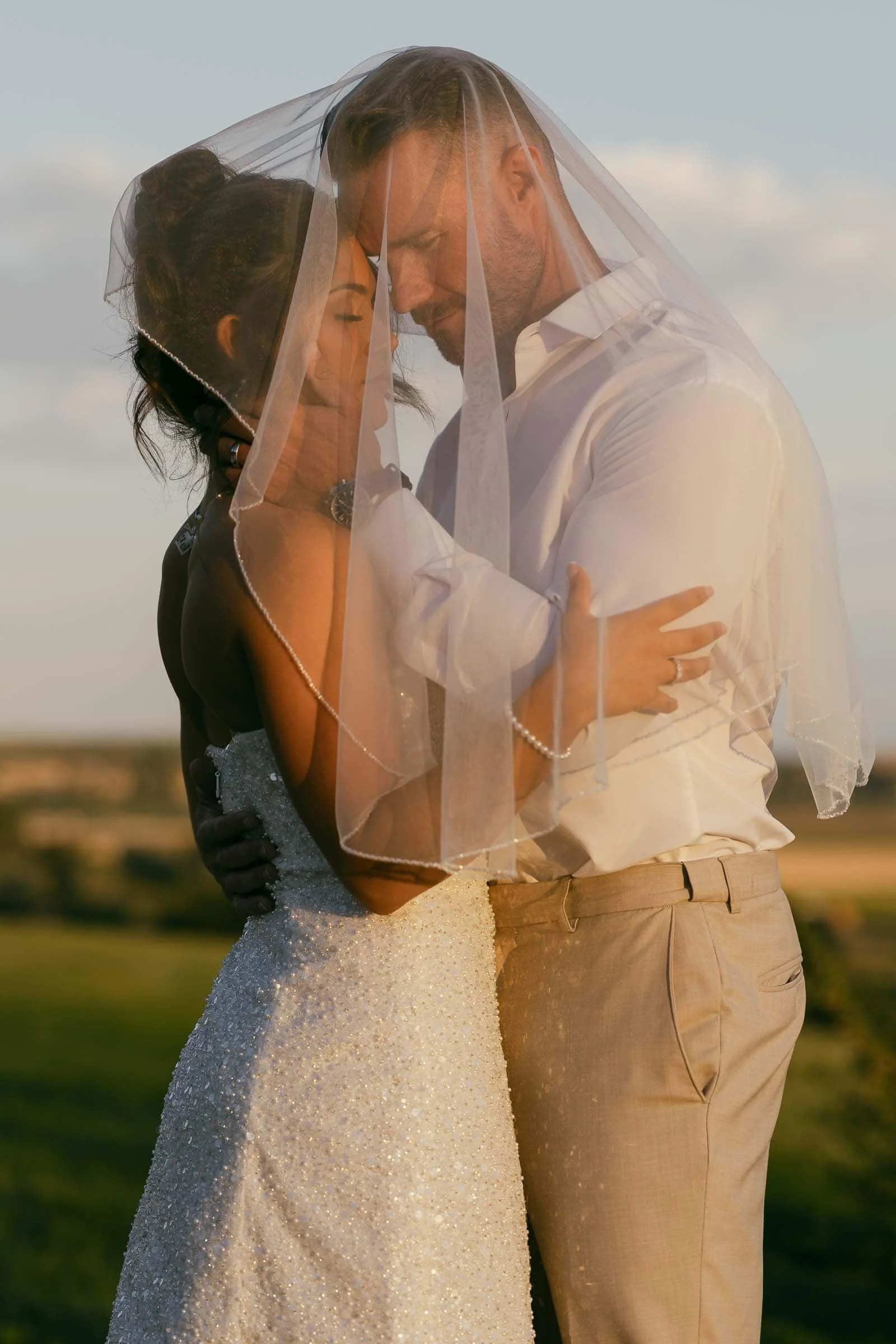  A bride and groom embrace closely outdoors at sunset, both under a sheer white veil. The bride wears a sparkly strapless dress, and the groom is in a white shirt and light trousers. They look serene and intimate. 