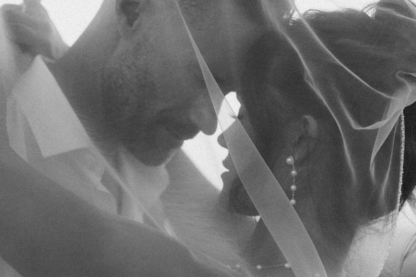  A black-and-white photo of a couple embracing closely, their faces touching gently under a sheer veil, creating a soft and intimate atmosphere. 