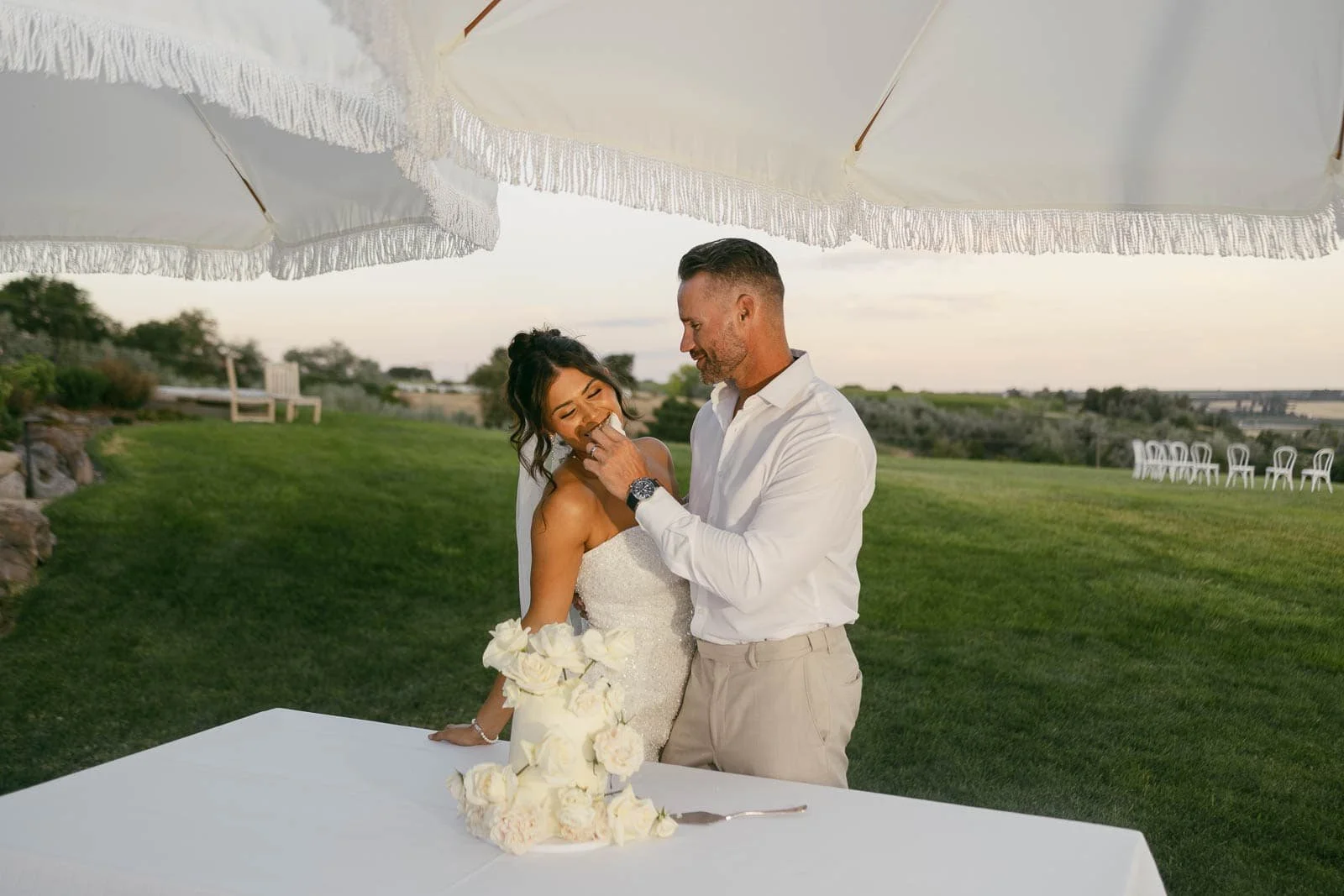  A bride and groom stand outdoors under white umbrellas, smiling at each other beside a white cake decorated with roses on a table, with green grass and chairs in the background. 