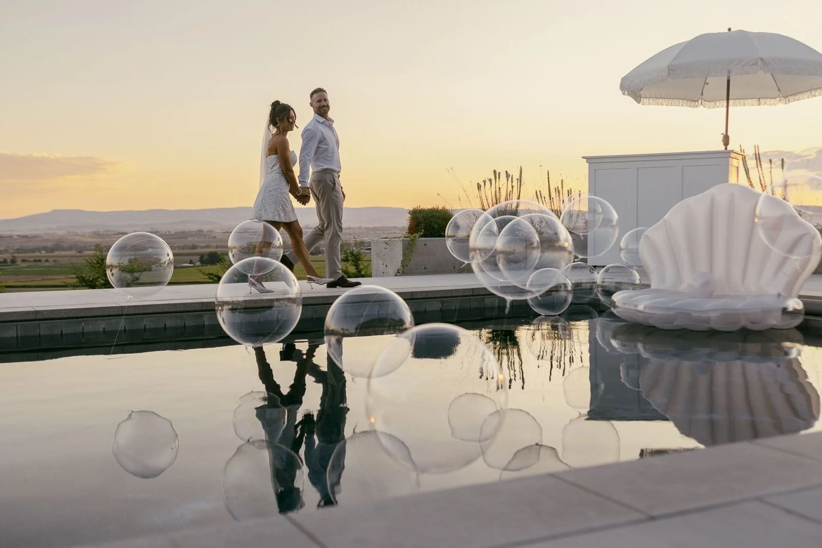  A couple holding hands walks along a pool at sunset, surrounded by transparent balloons. Their reflections are visible in the water. Nearby, there is a large clam-shaped seat and a white umbrella. The scene is elegant and romantic. 