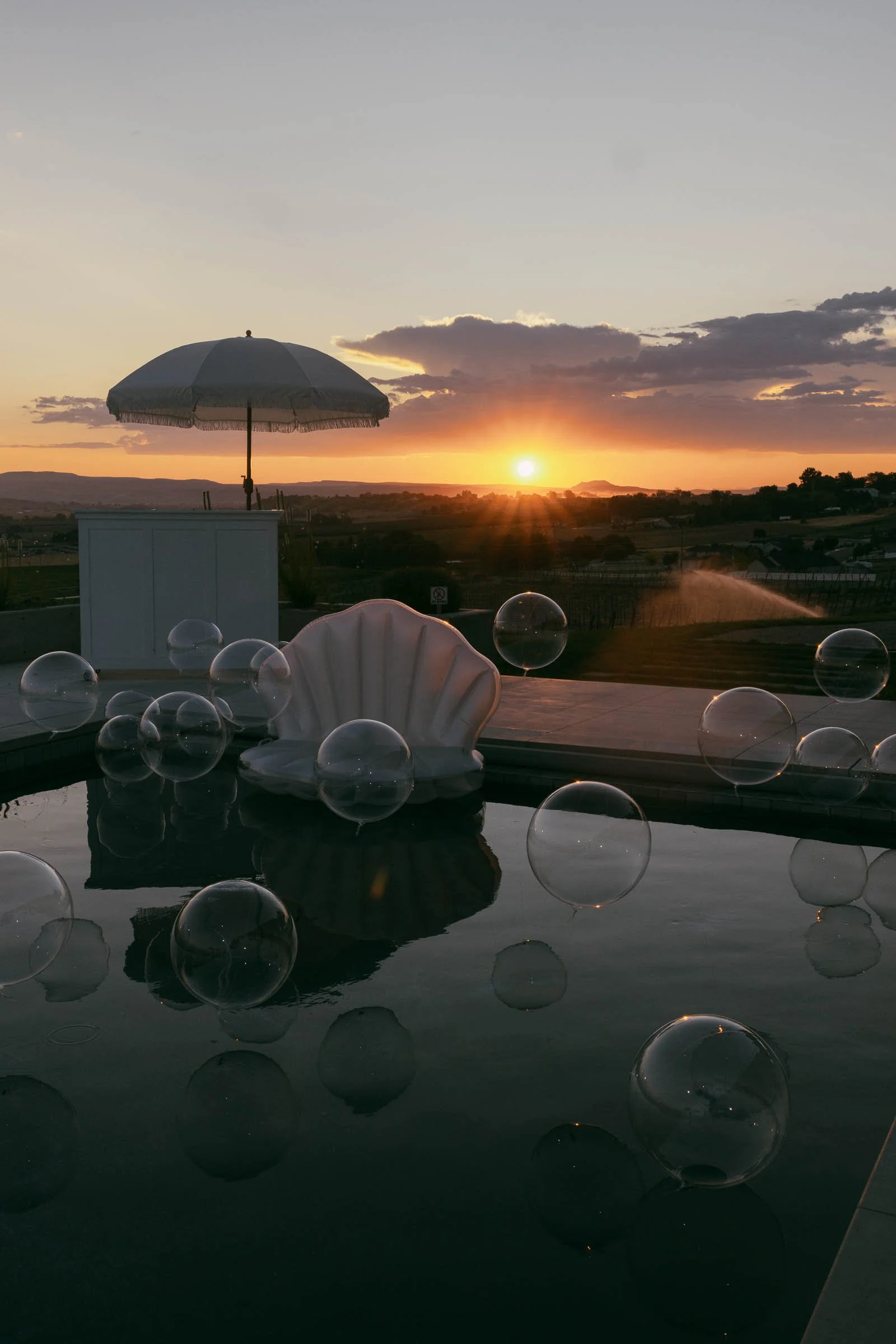 A sunset over a pool with transparent bubbles floating on the water, a large shell-shaped chair, and a white umbrella in the background. The sky is partly cloudy with warm orange and purple tones. 