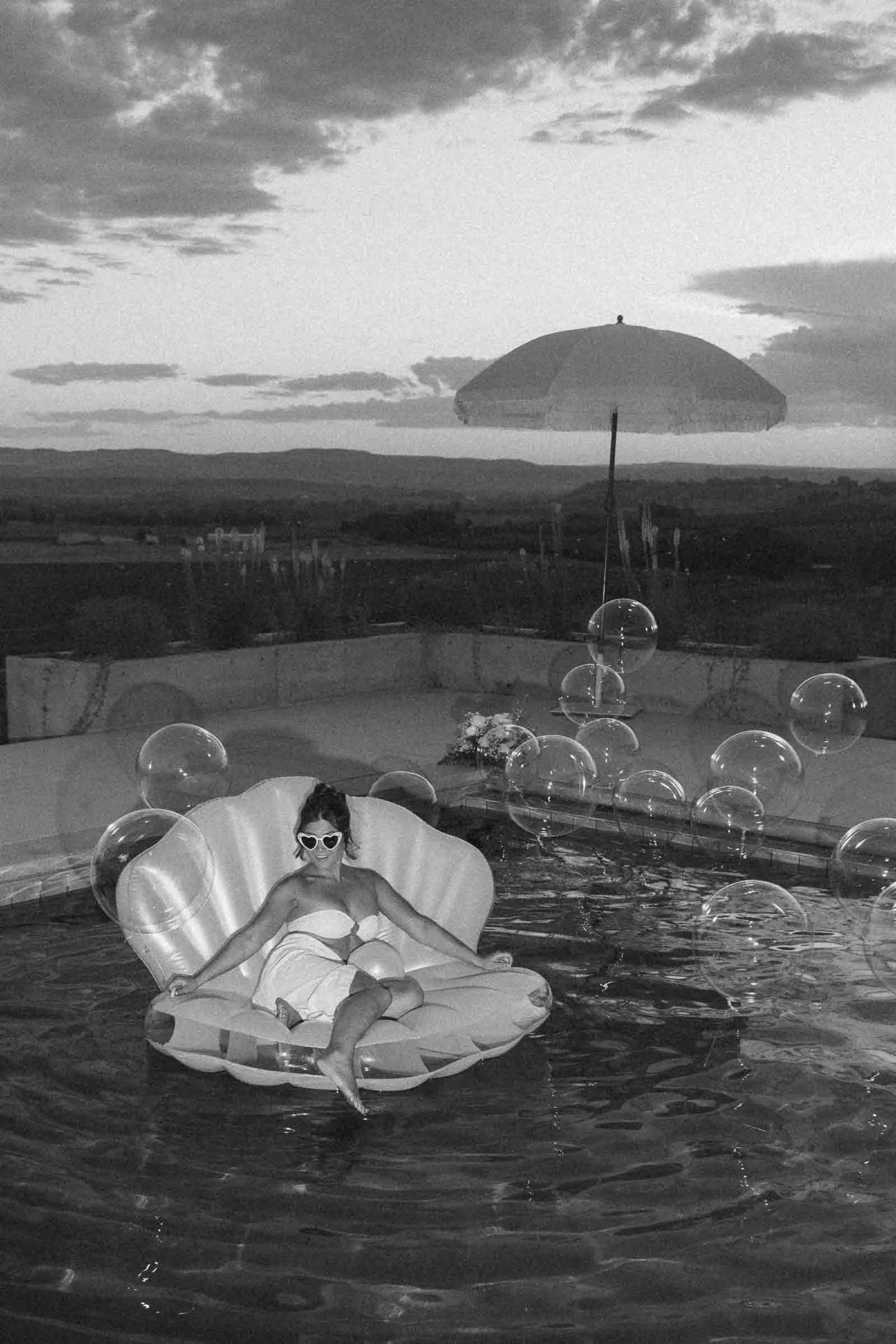  A woman wearing sunglasses and a towel sits on a shell-shaped pool float surrounded by bubbles. An umbrella stands beside the pool, with mountains and a cloudy sky in the background. The image is in black and white. 