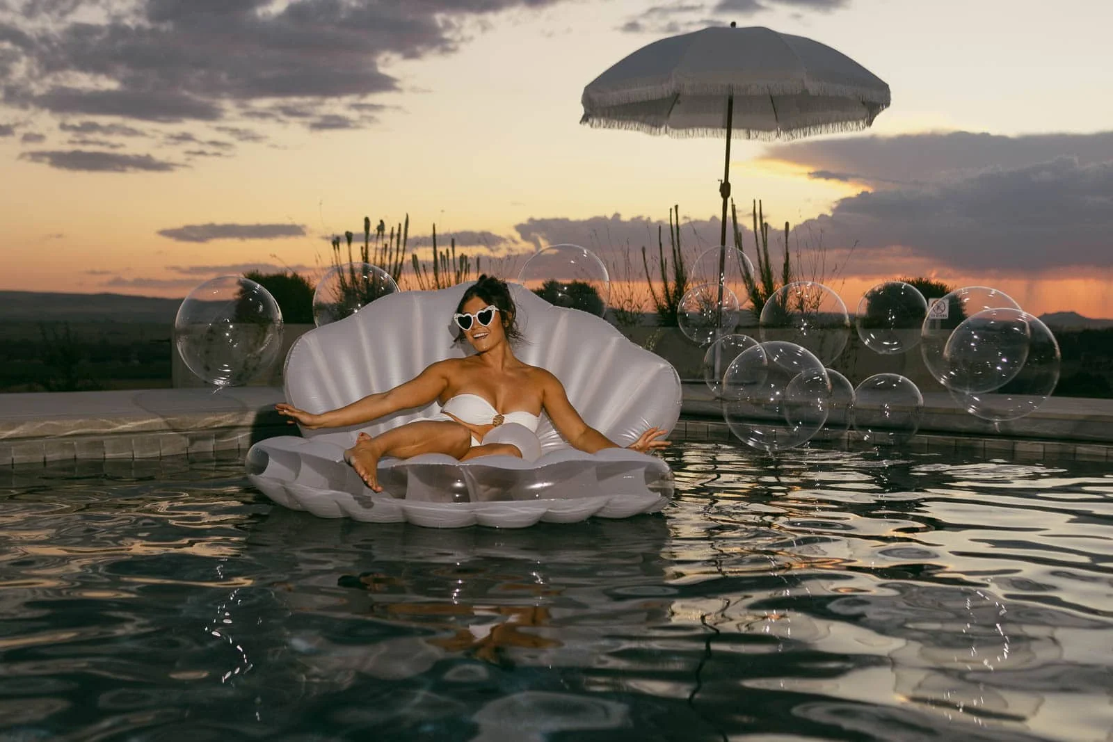  A woman in a white bikini and heart-shaped sunglasses relaxes on a clam-shaped pool float, surrounded by bubbles, with an umbrella and sunset in the background. 