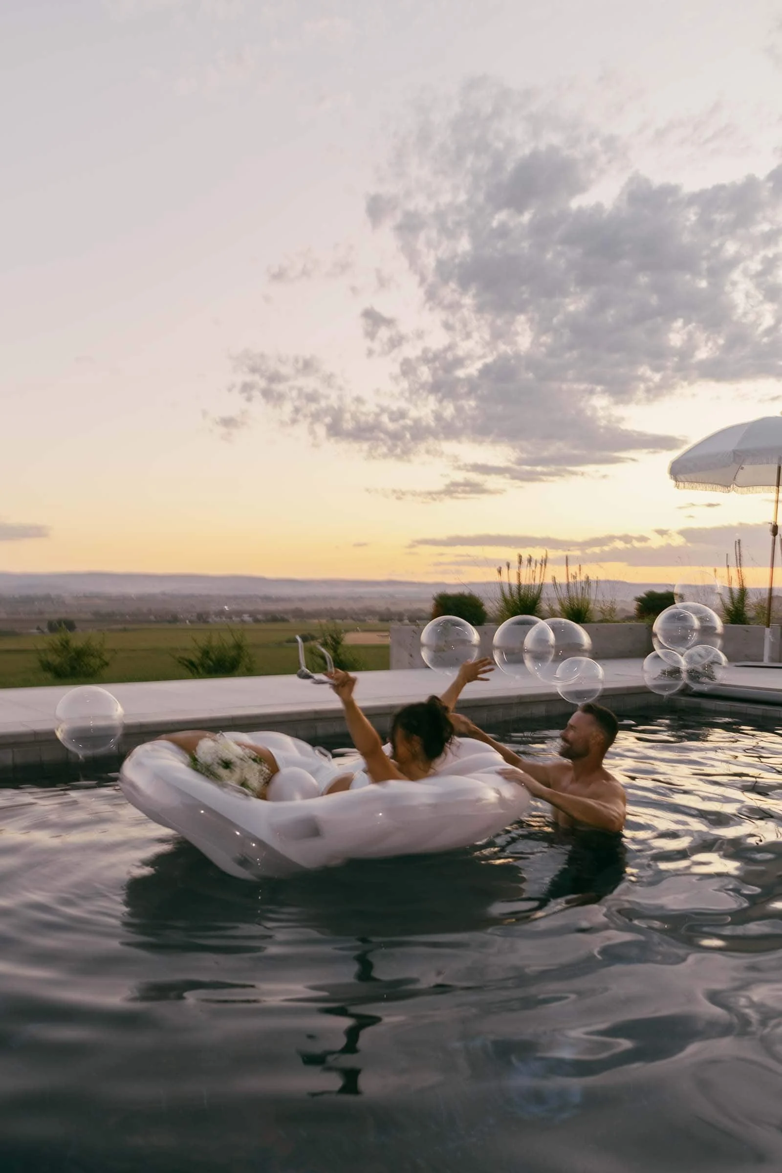  A man and woman relax in a pool at sunset, with the woman sitting on a shell-shaped float and the man in the water beside her. Large bubbles float around them, and an umbrella stands in the background. The image is in black and white. 