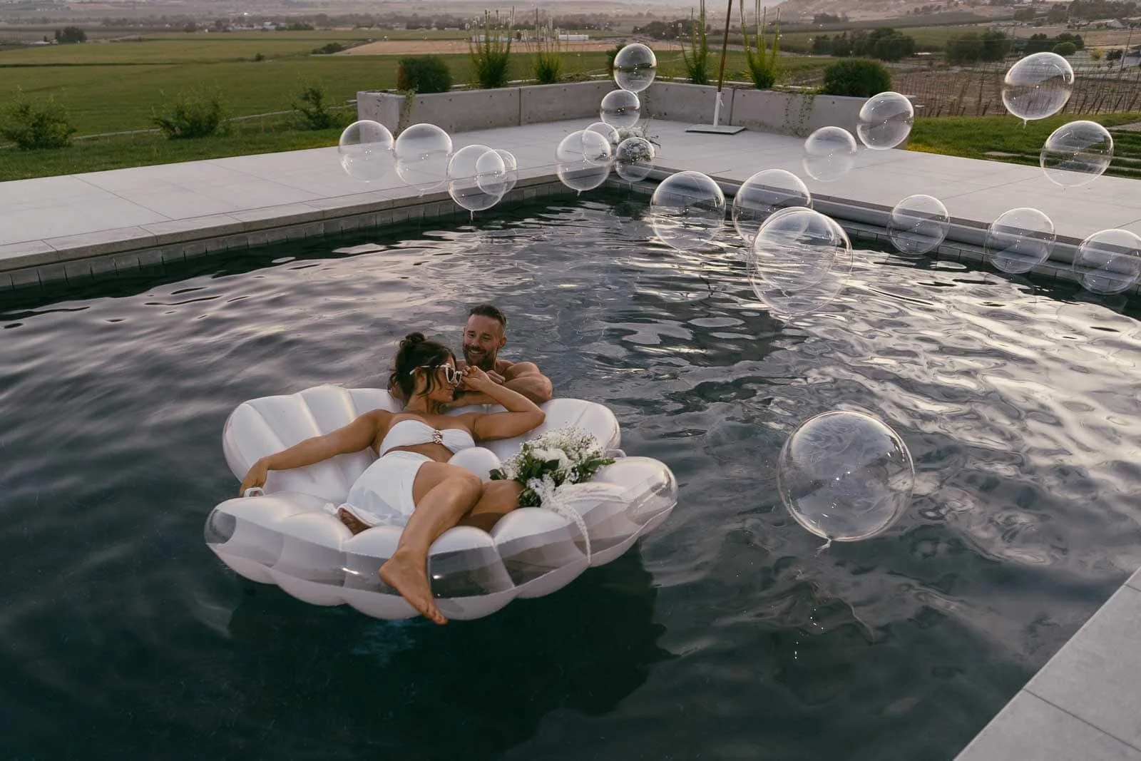A couple in wedding attire relaxes on a white pool float shaped like a shell in a pool, surrounded by floating bubbles, with a scenic landscape in the background.