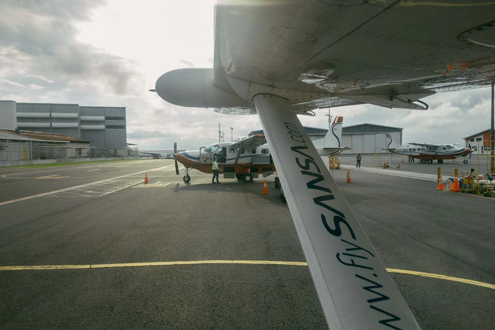  View from under an airplane wing showing several small aircraft parked on an airport tarmac under a cloudy sky, with hangars and a few people in the background. The wing displays the website " www.flySANSA.com ".  