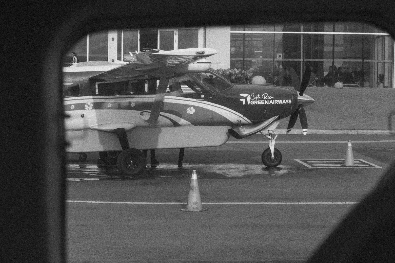  Black-and-white photo of a small propeller plane with "Green Airways" logo, parked on a wet tarmac near a building, viewed through a window frame with two traffic cones visible in front. 