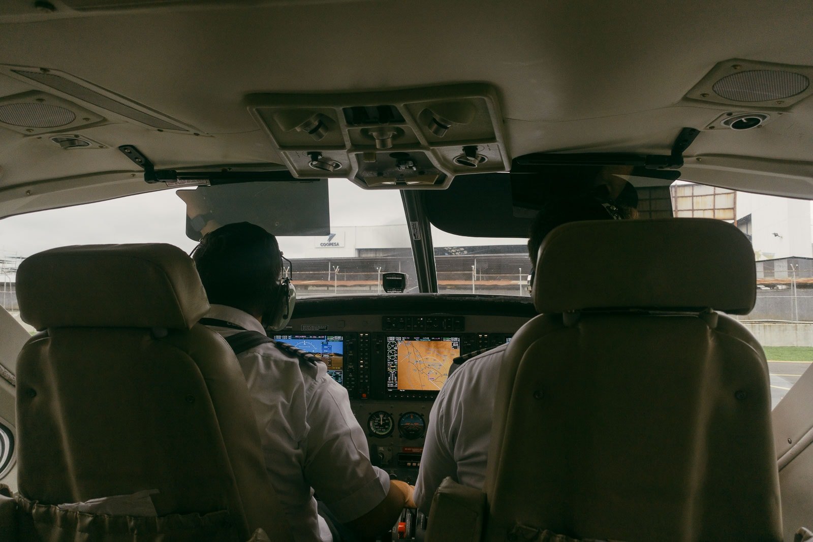  Two pilots in uniform sit inside a small aircraft cockpit, facing forward and preparing for takeoff, with flight instruments and navigation screens visible on the control panel. 
