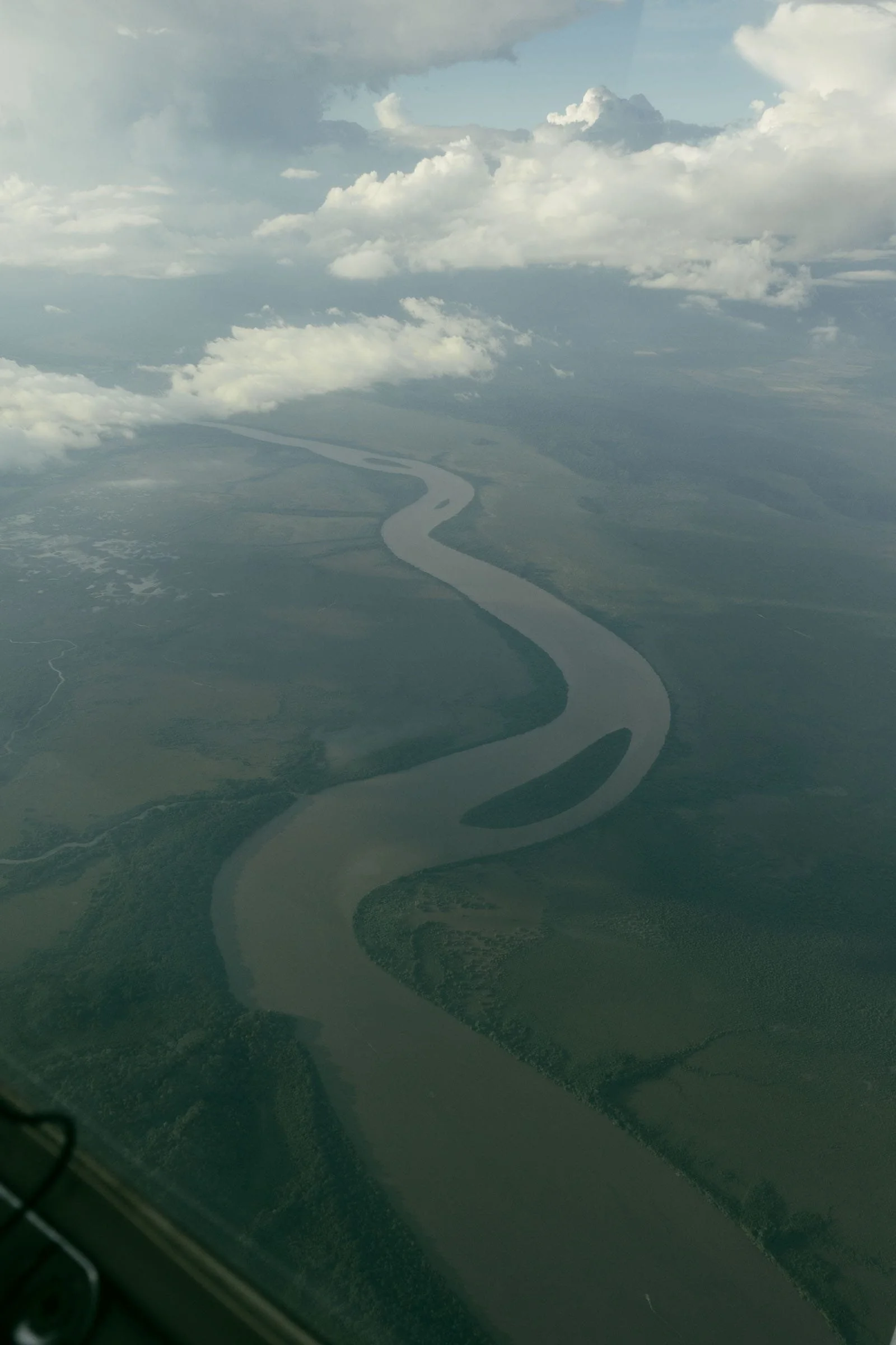  Aerial view of a winding river cutting through green landscape under a cloudy sky, with distant land and scattered patches of forest visible. 