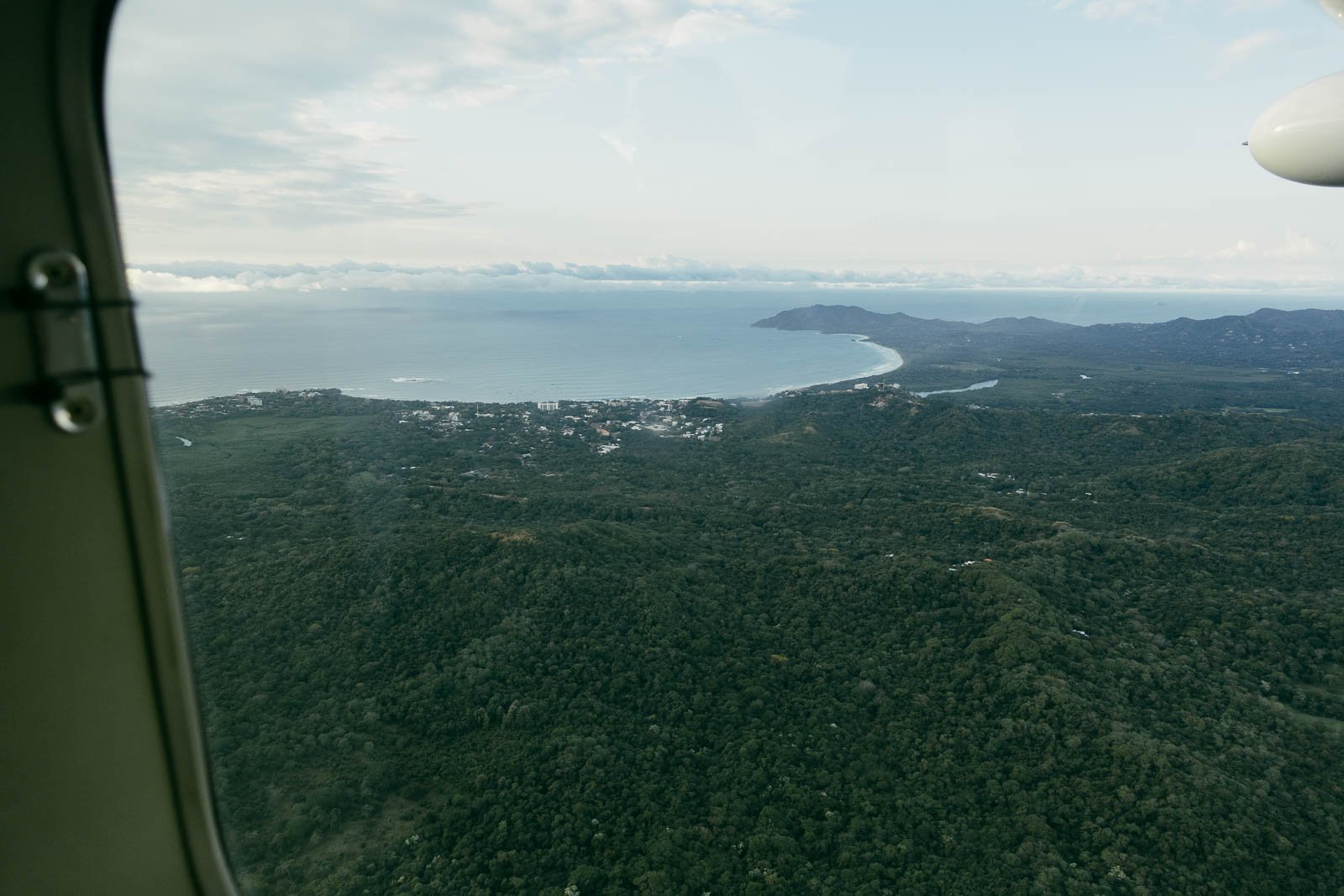  Aerial view from an airplane window overlooking a lush, green landscape, a coastline with beaches, and a town near the ocean under a partly cloudy sky. 