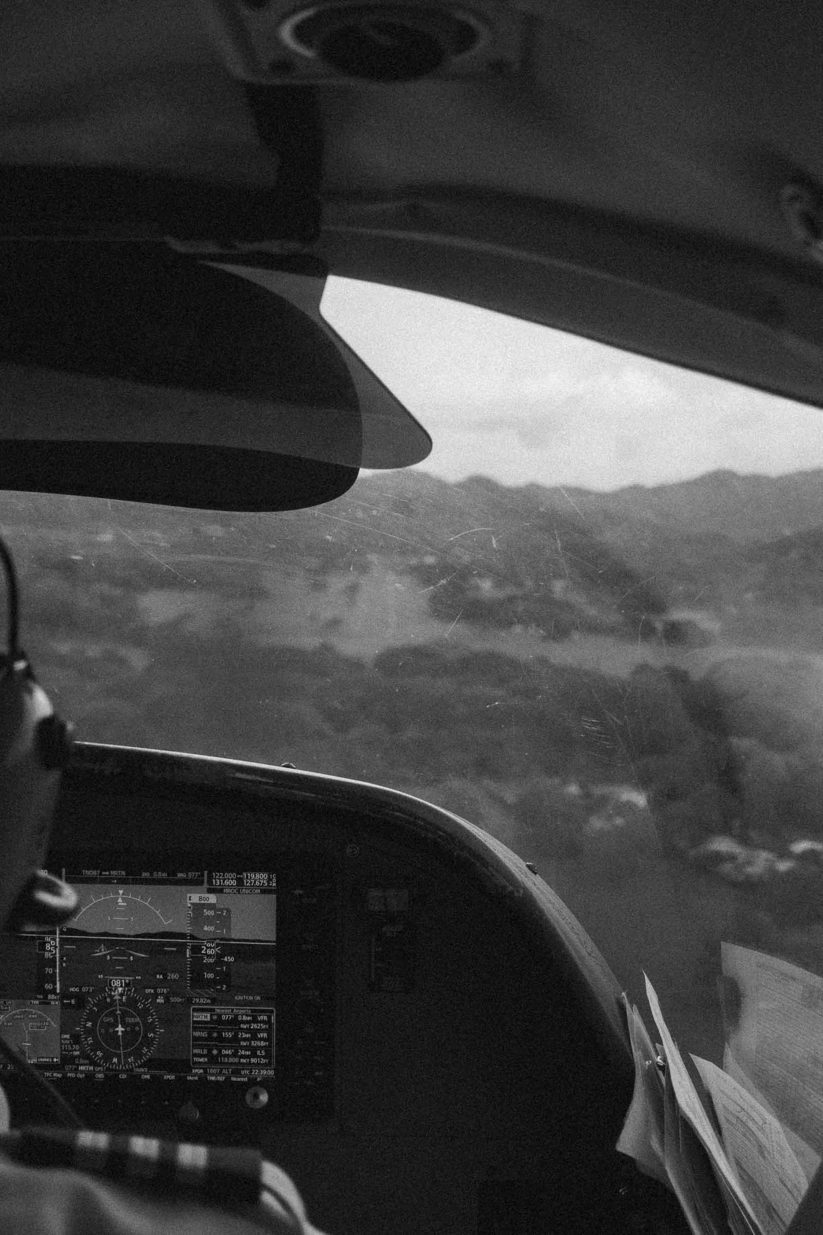  Black and white photo taken from behind a pilot inside a small aircraft, showing cockpit controls, part of the pilot’s head, and a view through the windshield of distant hills or mountains. 