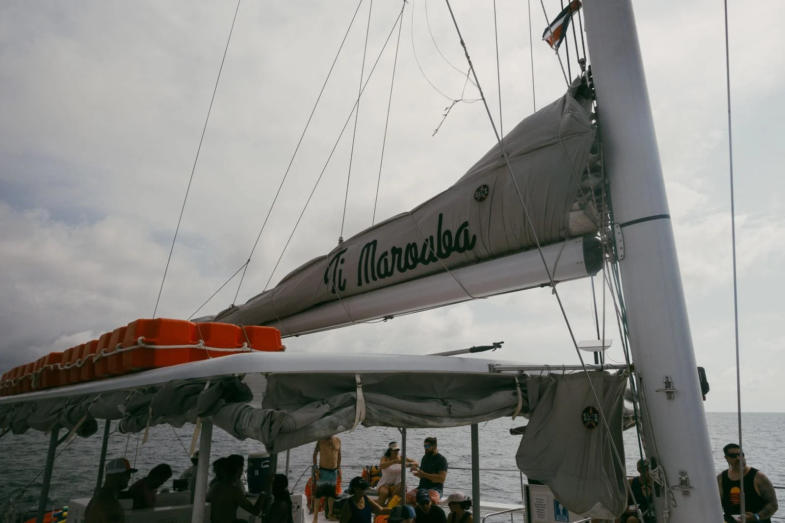  A group of people stand and sit on a sailboat named "La Marsalada" out on the ocean, under a cloudy sky. Some wear swimwear and hats. Orange life vests are stored above the boat’s shaded area. 