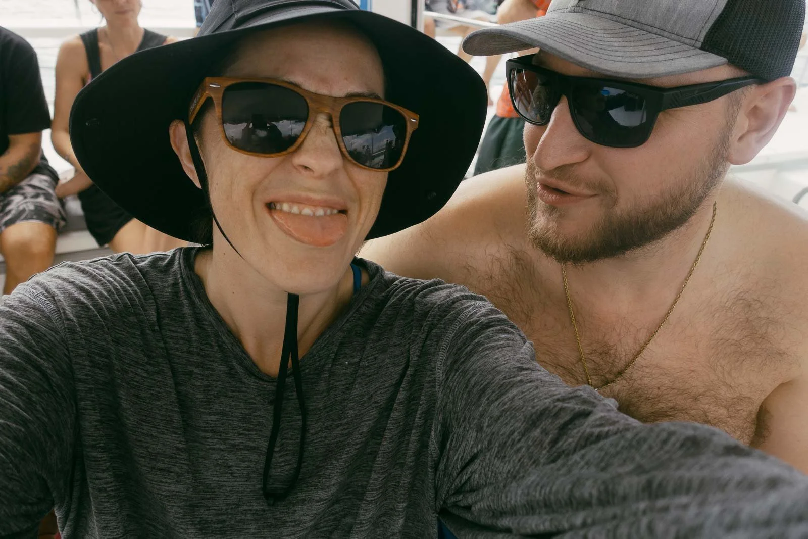  Two people taking a selfie on a boat; one person is wearing sunglasses, a wide-brim hat, and sticking out their tongue, while the other is shirtless, wearing sunglasses and a cap, smiling slightly. Others are in the background. 