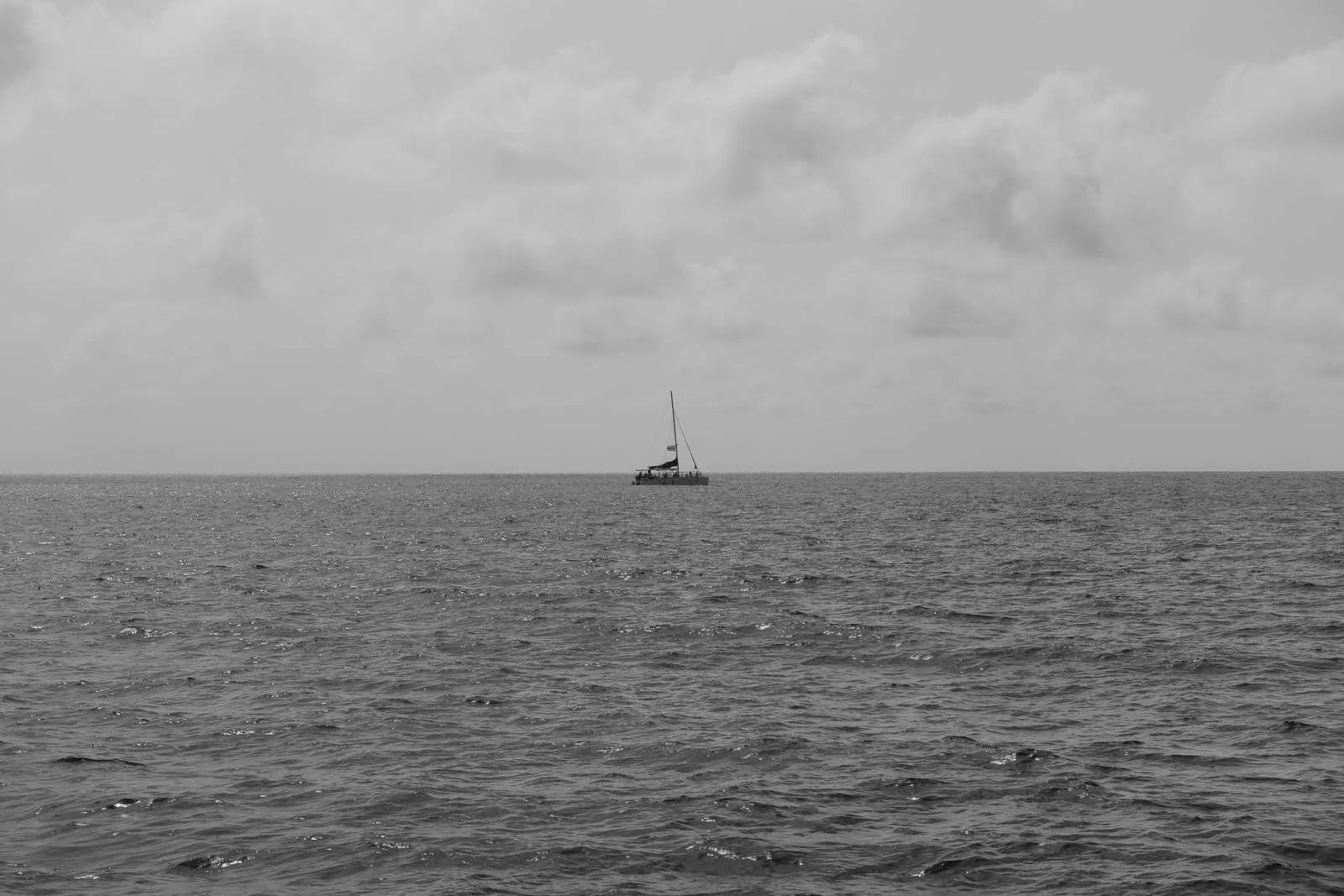  A sailboat is seen in the distance on a vast, calm ocean under a cloudy sky. The image is in black and white, with gentle waves in the foreground. 