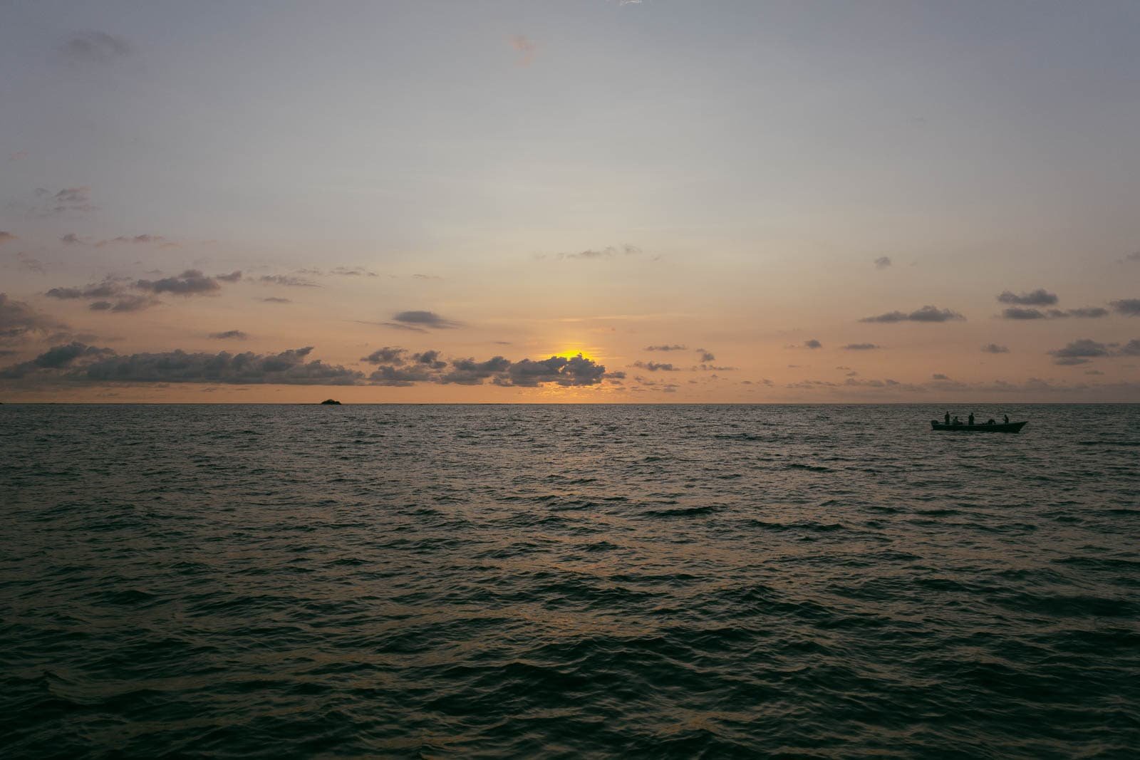  A calm ocean at sunset with gentle waves, a small boat with people is visible on the right, and scattered clouds are in the sky. 