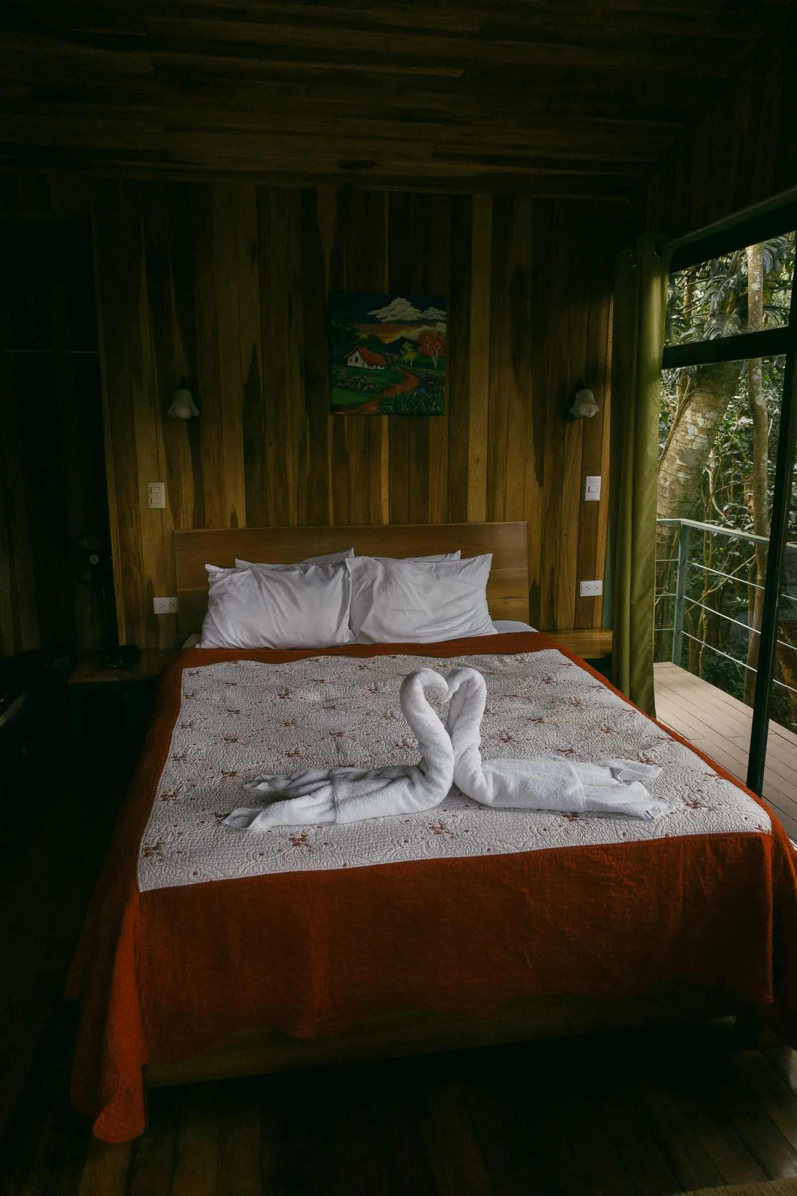  A cozy bedroom with wooden walls, a bed covered in a white and orange blanket, white pillows, and two towel swans forming a heart shape. A painting hangs above the bed, and a glass door opens to a balcony with trees outside. 