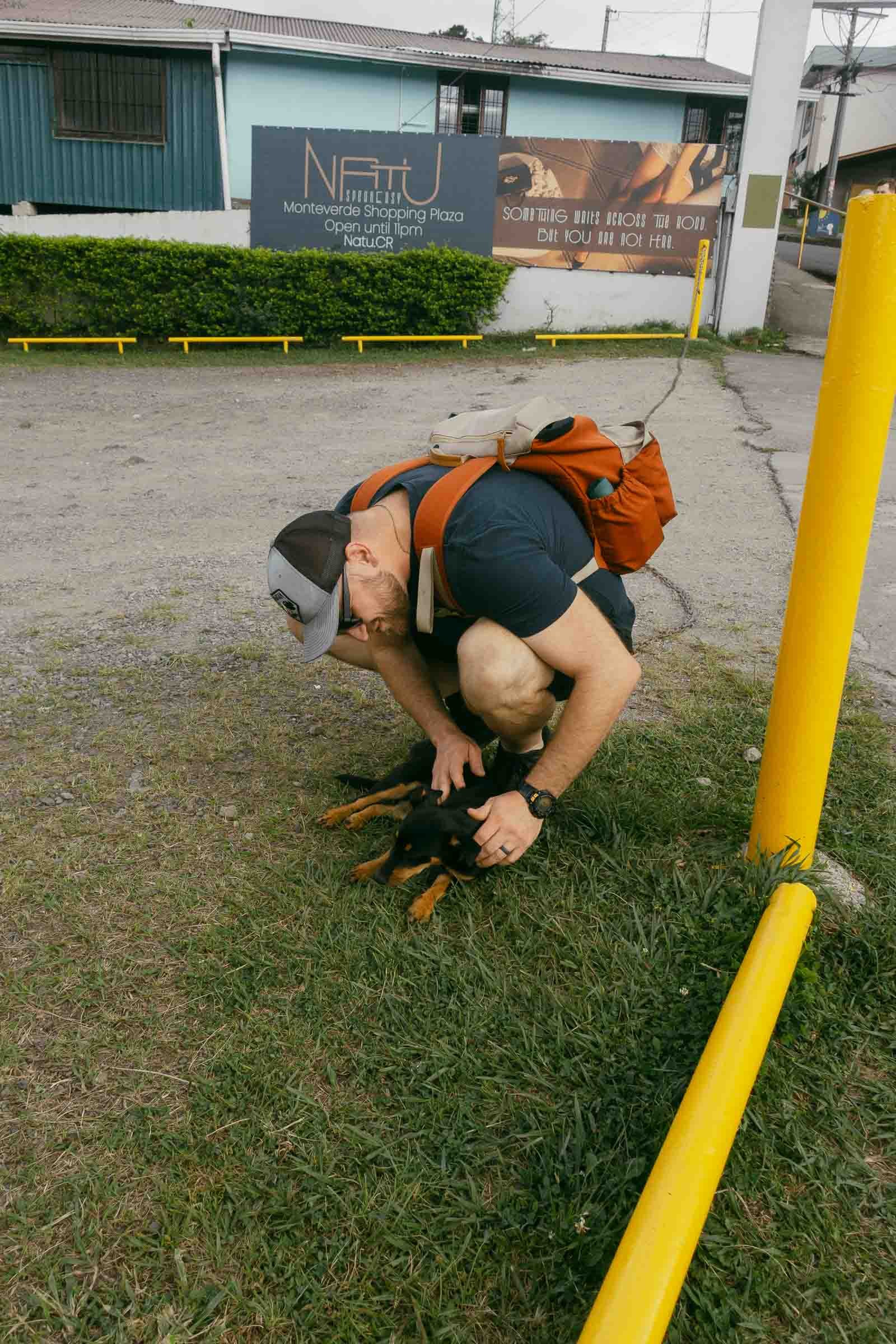  A man wearing a cap, glasses, and a backpack crouches on grass, petting a small black dog near a yellow pole in front of a building with signs and greenery. 