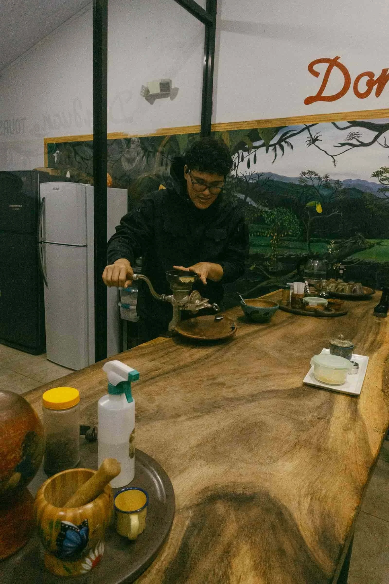  A person stands behind a wooden counter using a manual coffee grinder. The counter holds various containers and utensils. A refrigerator and a mural with greenery and the word "Don" are seen in the background. 