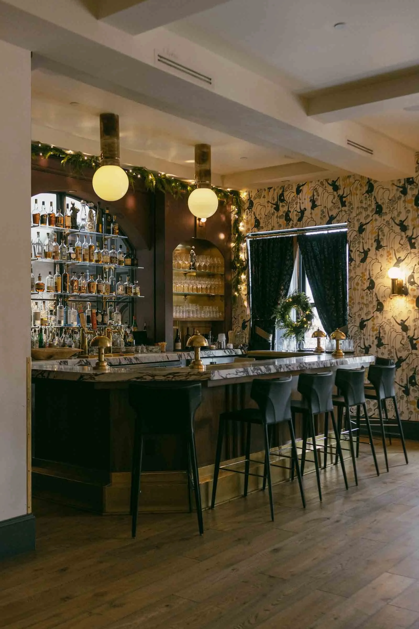  A stylish bar with a marble countertop, black chairs, and shelves filled with bottles. Warm lighting, decorative garland, patterned wallpaper, and a window with dark curtains create a cozy atmosphere. The floor is wooden. 