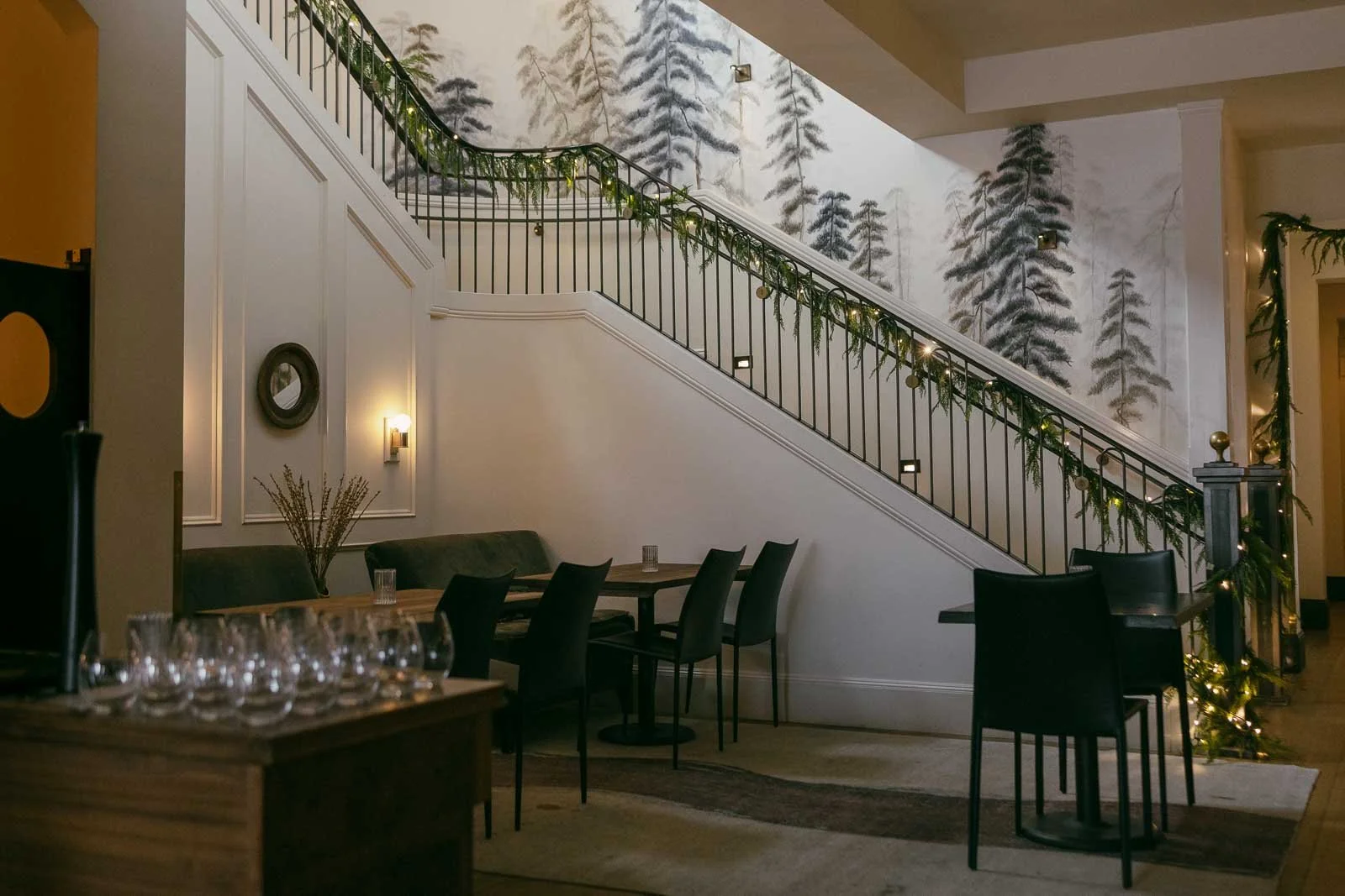  A modern dining area with black chairs and tables, a staircase decorated with garland, and a wall mural of pine trees in the background. Wine glasses are arranged on a wooden table in the foreground. 