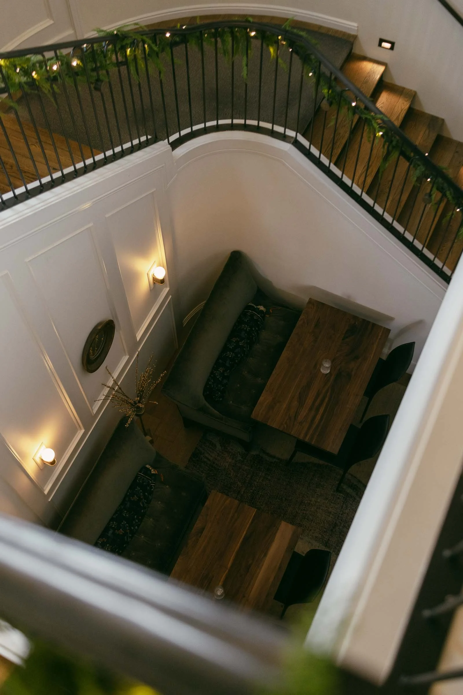  A top-down view of a cozy seating area with two wooden tables and green cushioned benches, located beneath a curved staircase decorated with garland and string lights. 