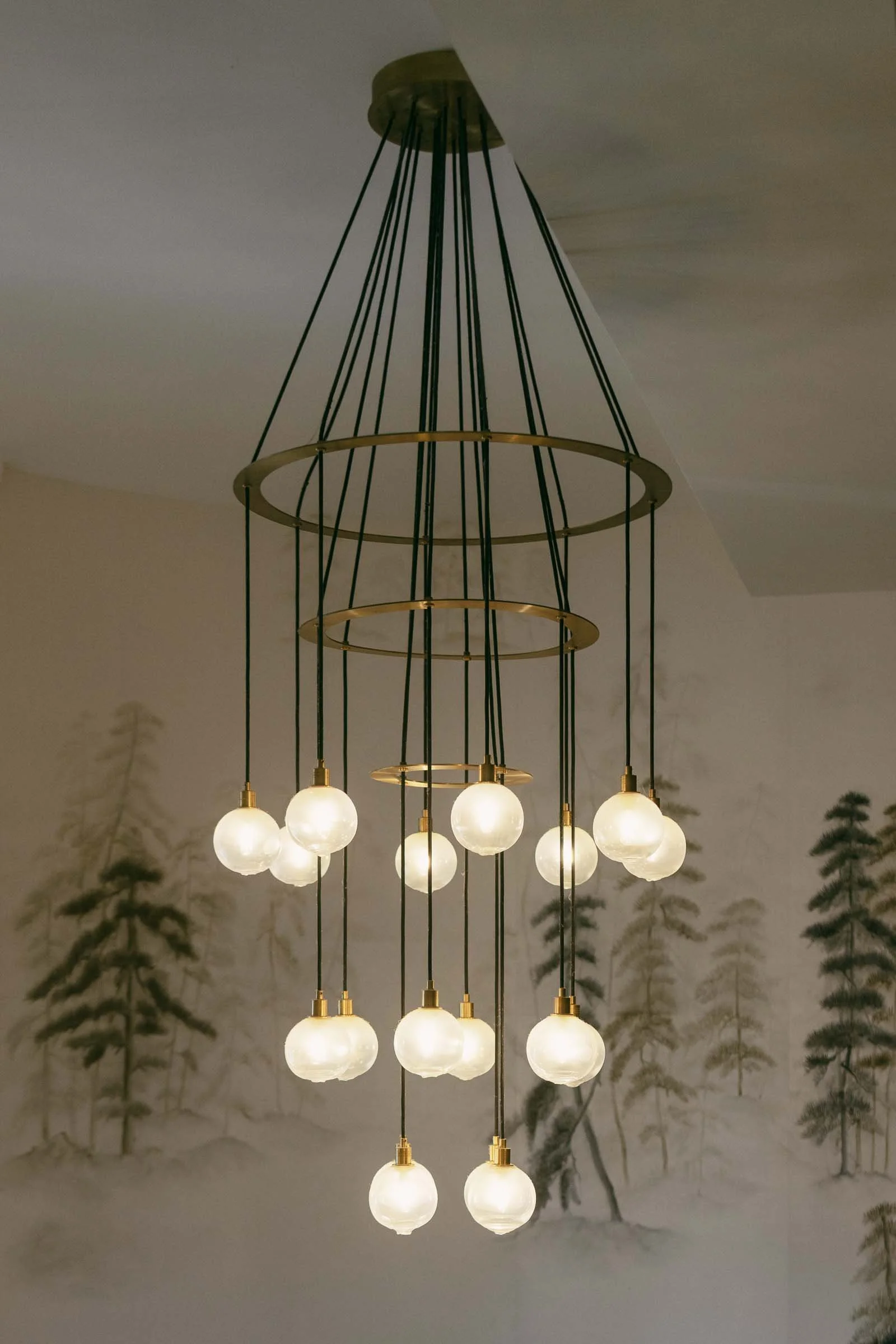  A modern chandelier with two circular metal frames and twelve hanging glass globes emits warm light. The chandelier is suspended from the ceiling in a room with a wall featuring a subtle tree mural. 