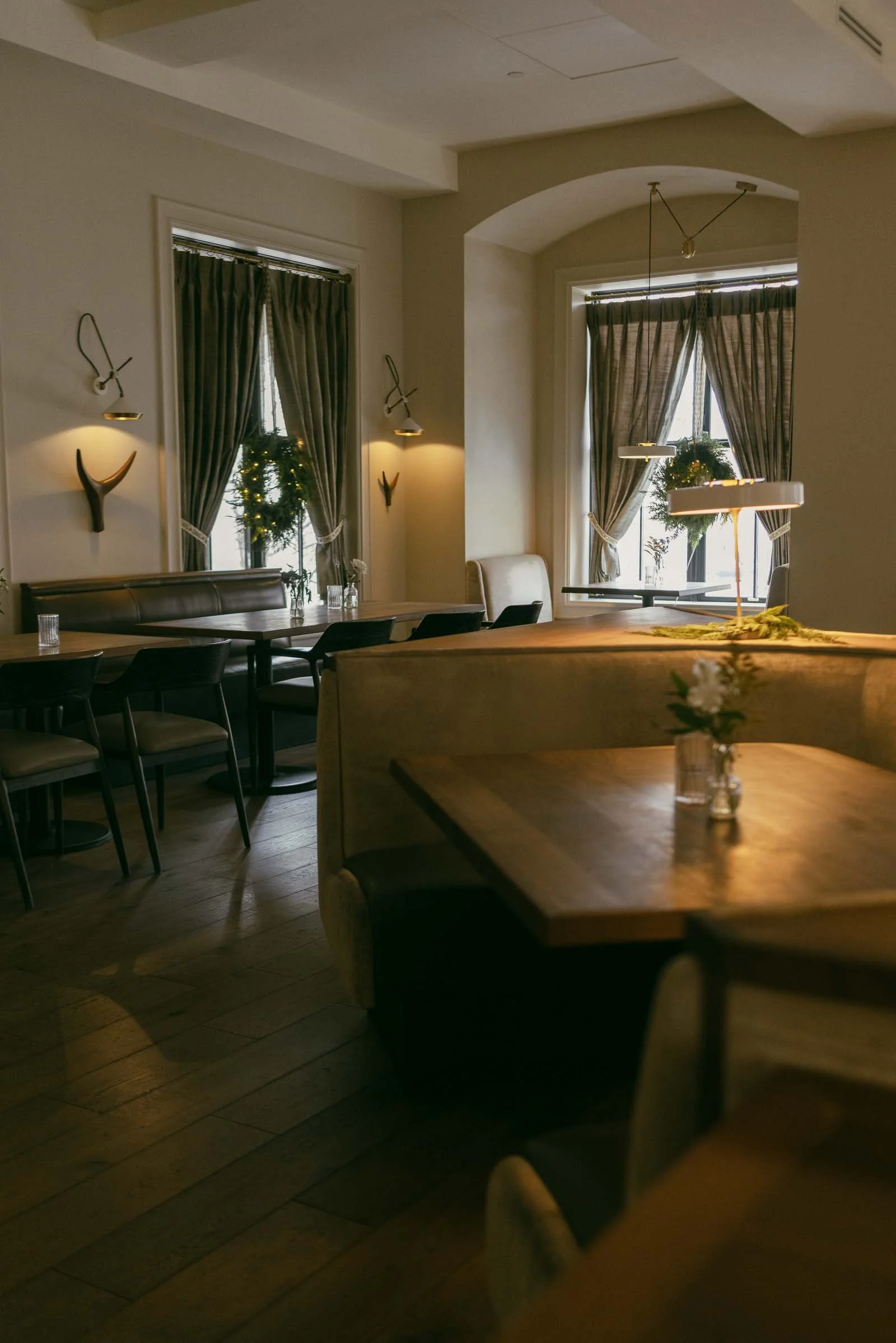  A warmly lit restaurant interior with wooden tables, beige upholstered booths, and chairs. Sunlight filters through curtains on two windows, and small vases with flowers decorate the tables. 