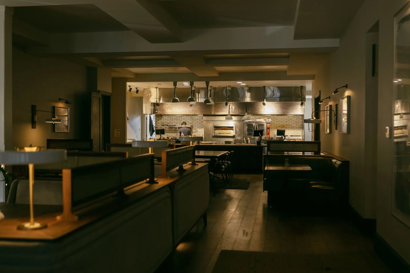  A dimly lit, modern restaurant interior with empty booths and tables, wooden floors, and an open kitchen in the background where a chef is working. Warm lights create a cozy atmosphere. 