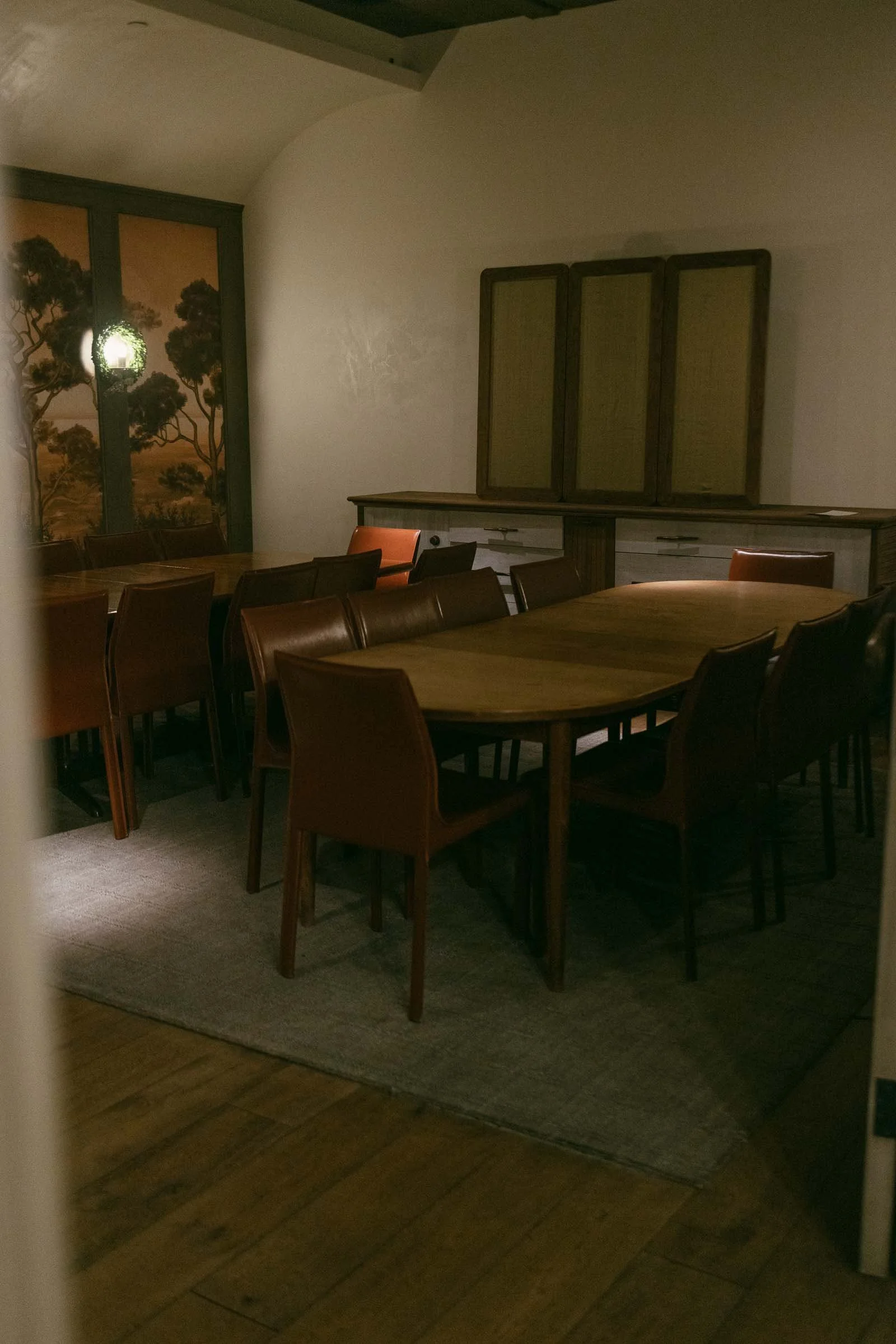  A dimly lit private dining room with two wooden tables, surrounded by brown leather chairs on a gray rug. A cabinet and wall art featuring trees are visible in the background. The room has wooden floors and an arched ceiling. 