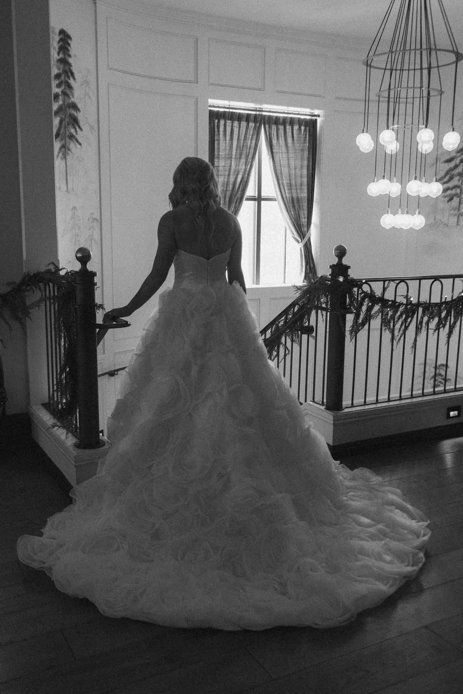  A woman in a strapless, full-skirted wedding gown stands on a staircase, facing away from the camera. Soft light shines through a window ahead, highlighting the intricate details of her dress. 