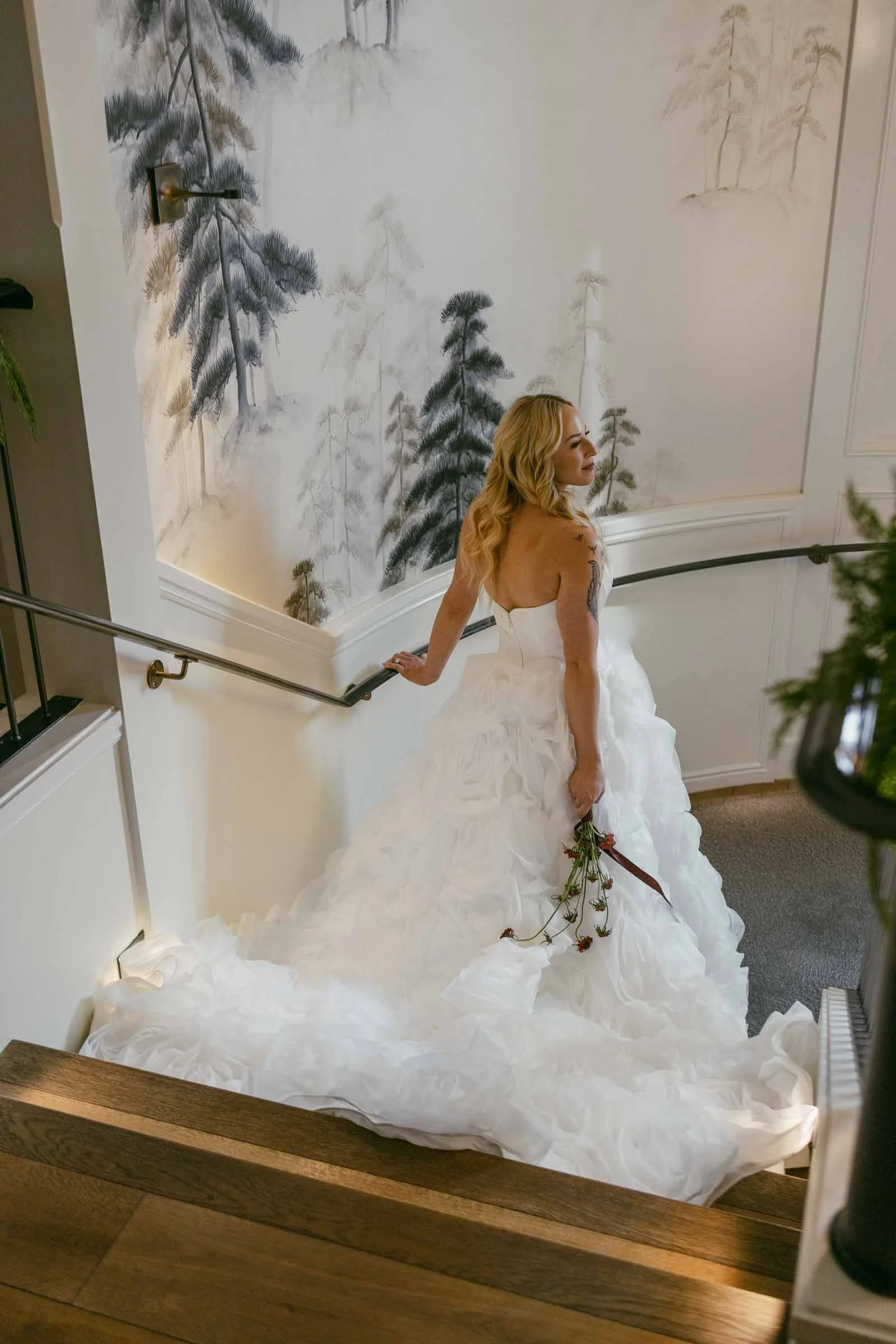  A bride in a strapless white gown holds dried red roses while standing on a staircase, looking over her shoulder. The wall behind her is decorated with soft, painted trees. 