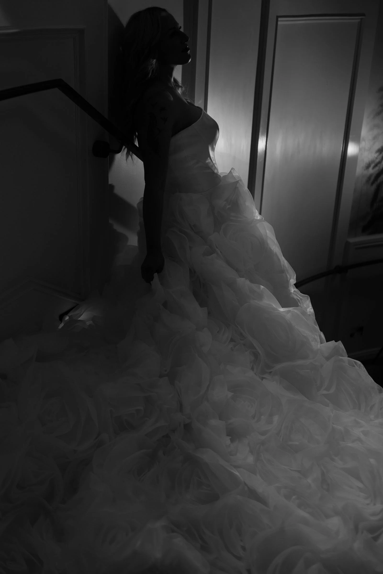  A woman in a voluminous, ruffled gown stands on a staircase, leaning against the wall. Soft light highlights the texture of her dress, creating dramatic shadows in the black and white scene. 