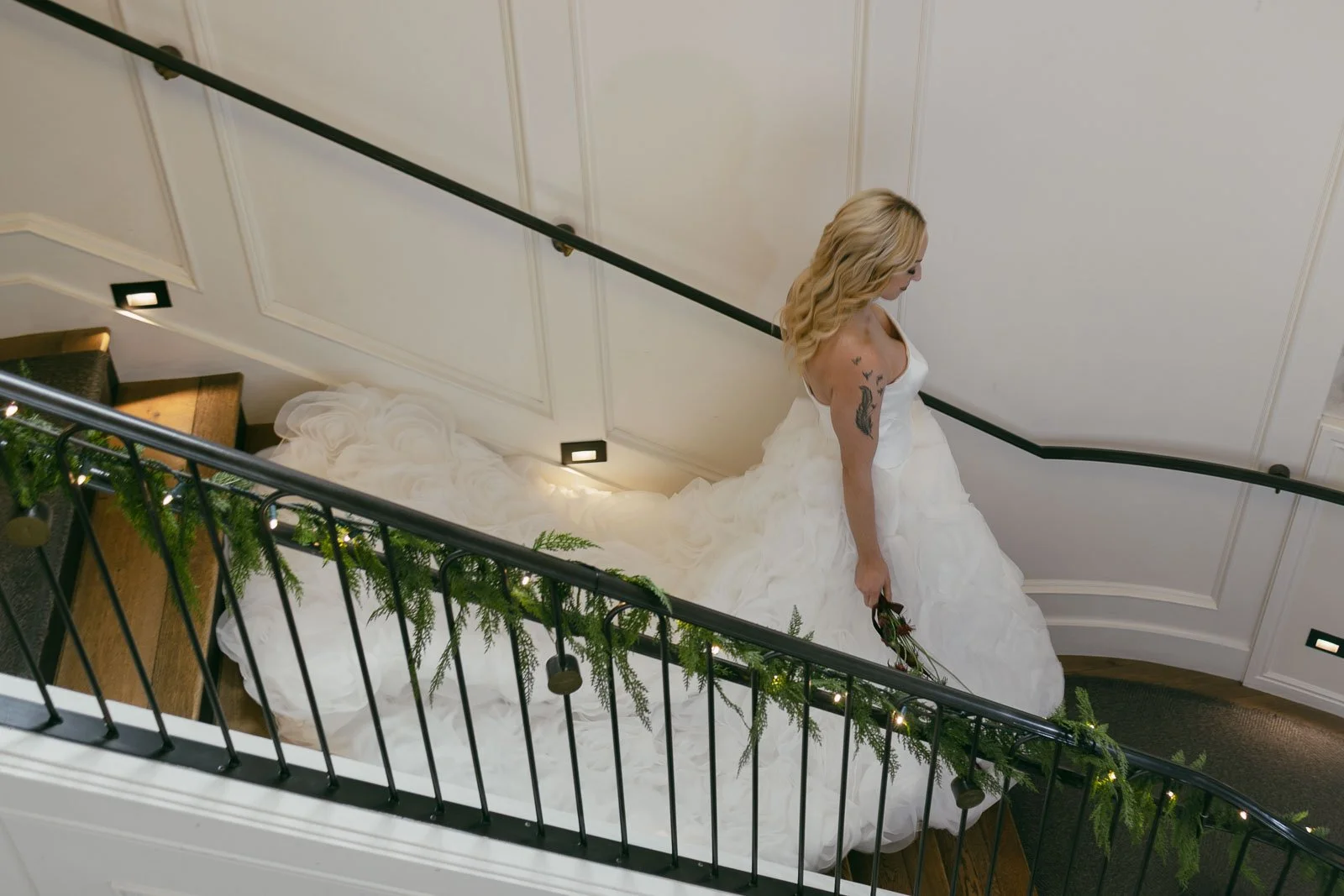 A bride in a white gown with a long train walks down a carpeted staircase decorated with greenery, holding a bouquet and looking downwards. The wall beside her is adorned with elegant paneling.