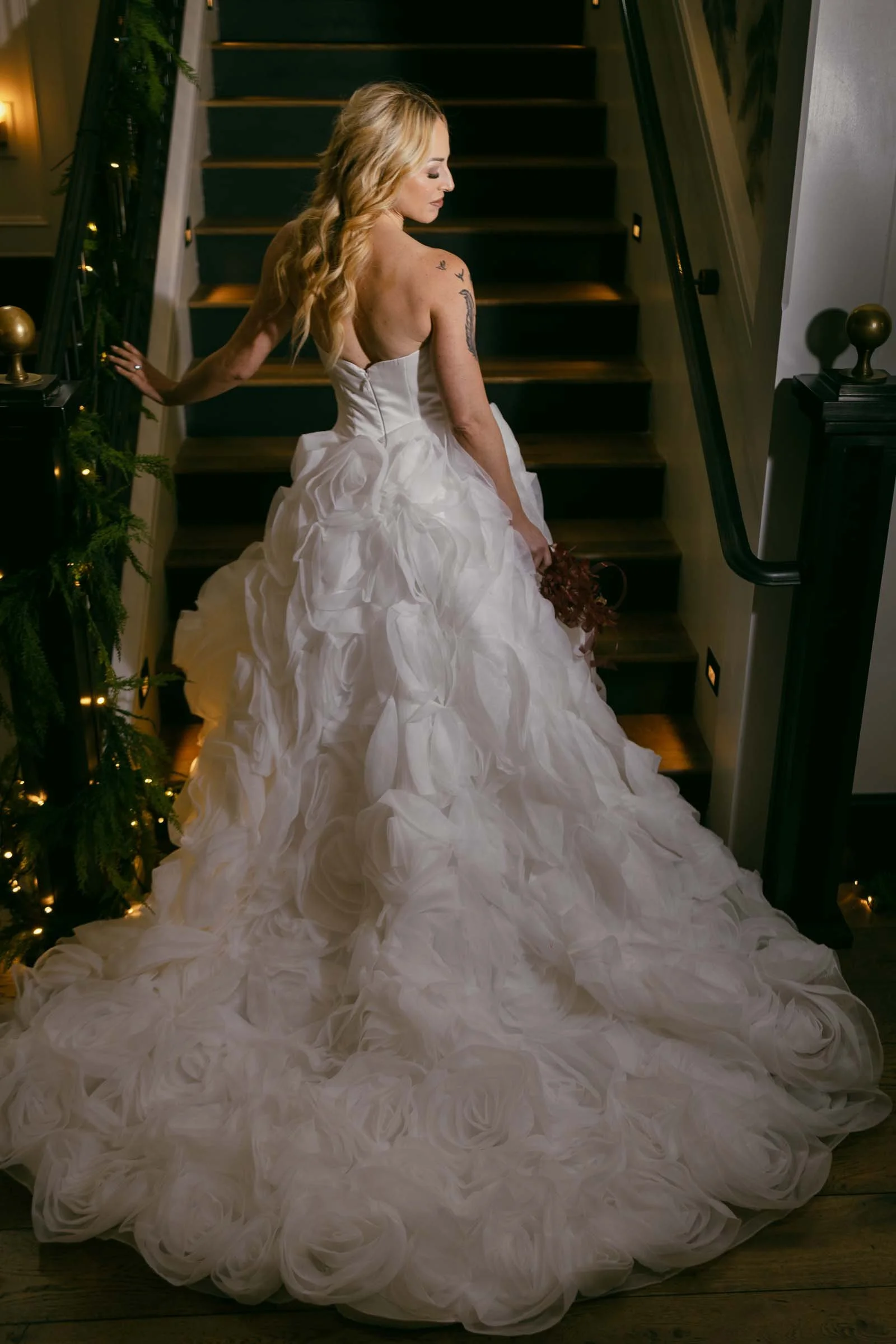  A woman in a strapless, flowing white wedding dress with a long, ruffled train stands on stairs, looking over her shoulder. She holds a bouquet and has long, wavy blonde hair. The scene is softly lit and decorated with greenery. 