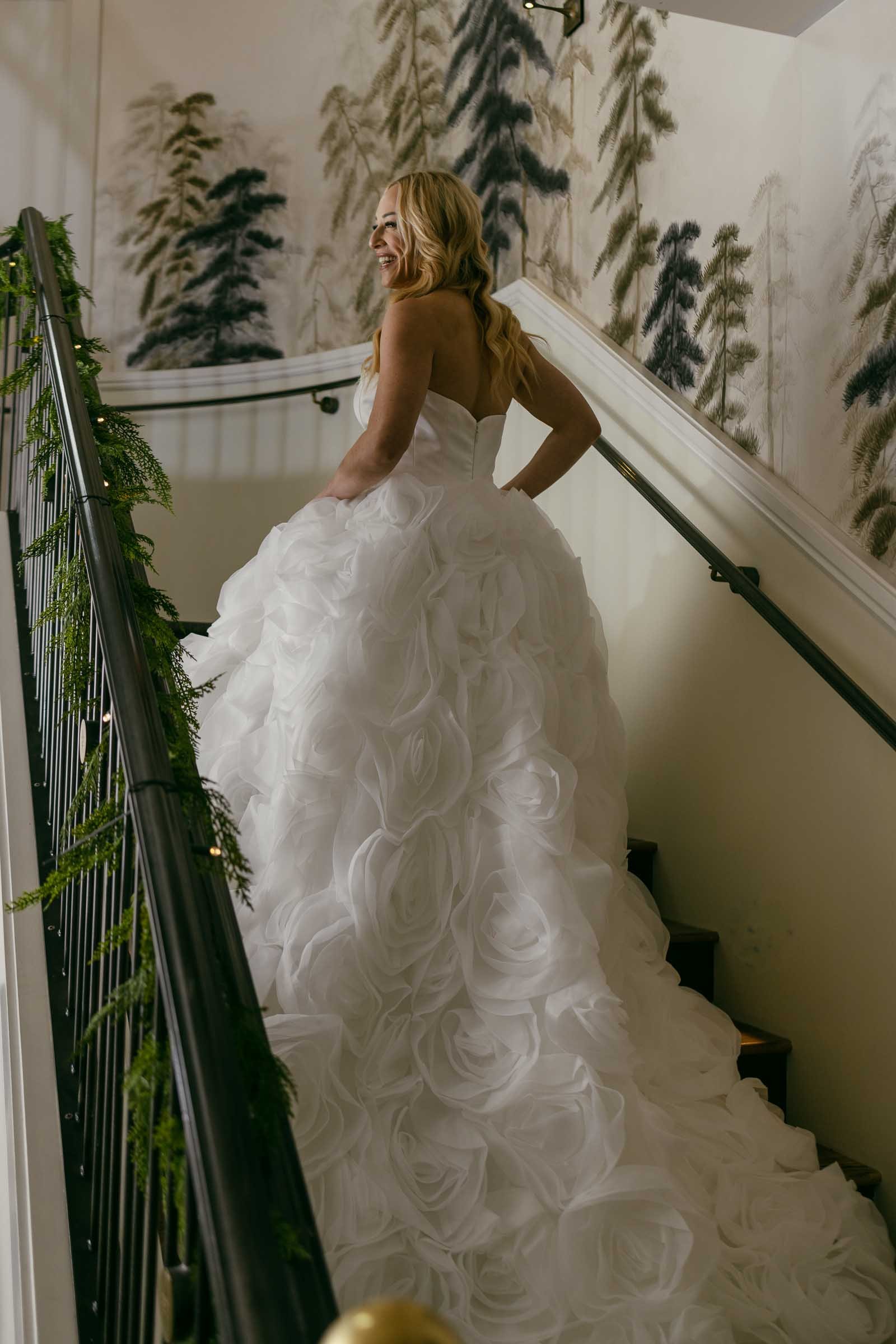 A woman in a strapless white wedding gown with a voluminous, rose-patterned skirt stands on a staircase decorated with greenery, looking over her shoulder and smiling. Forest-themed wallpaper lines the wall behind her.