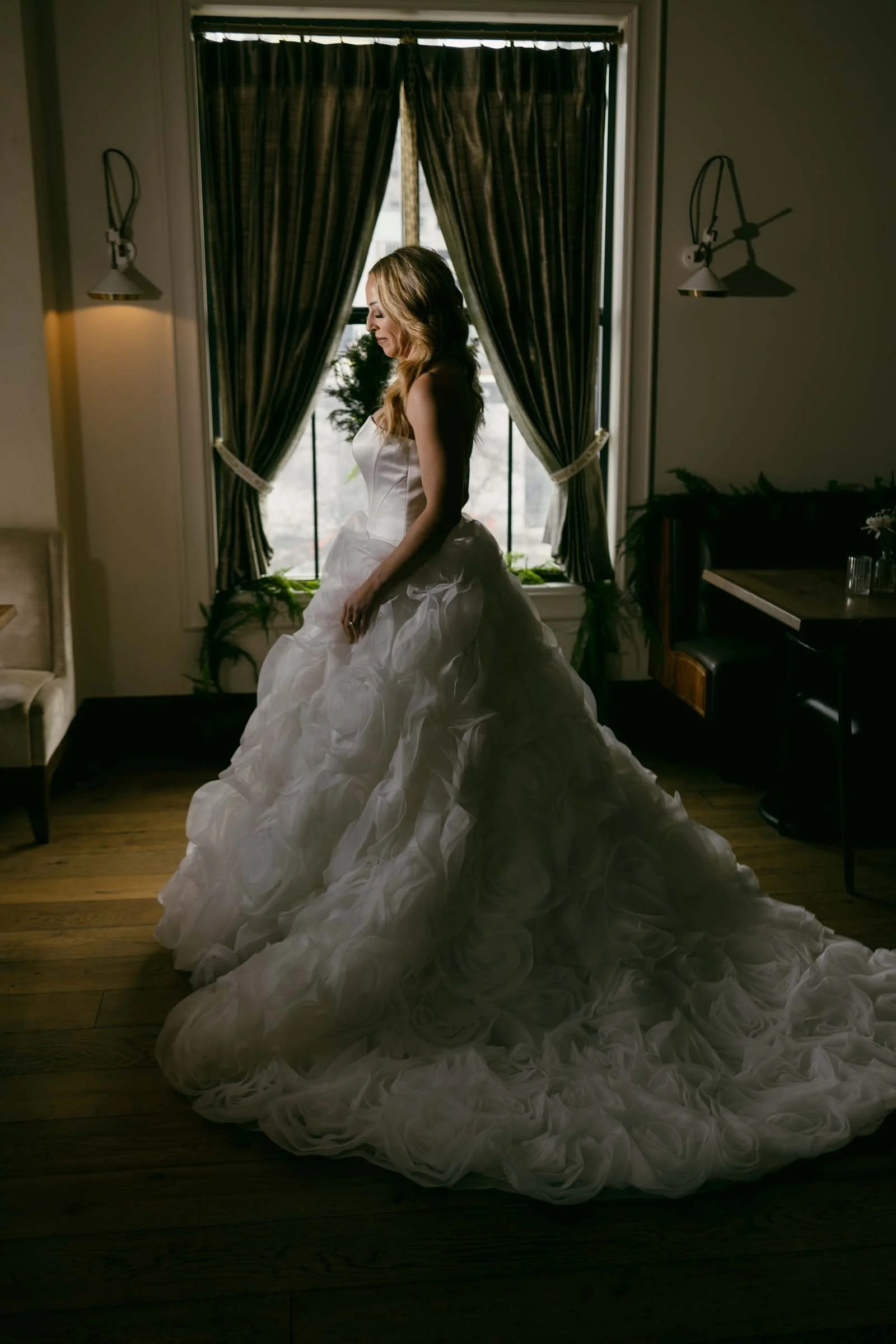A woman in a white, textured, voluminous wedding dress stands in a dimly lit room with wooden floors and tall windows framed by dark curtains. Light from the window highlights her profile and gown.