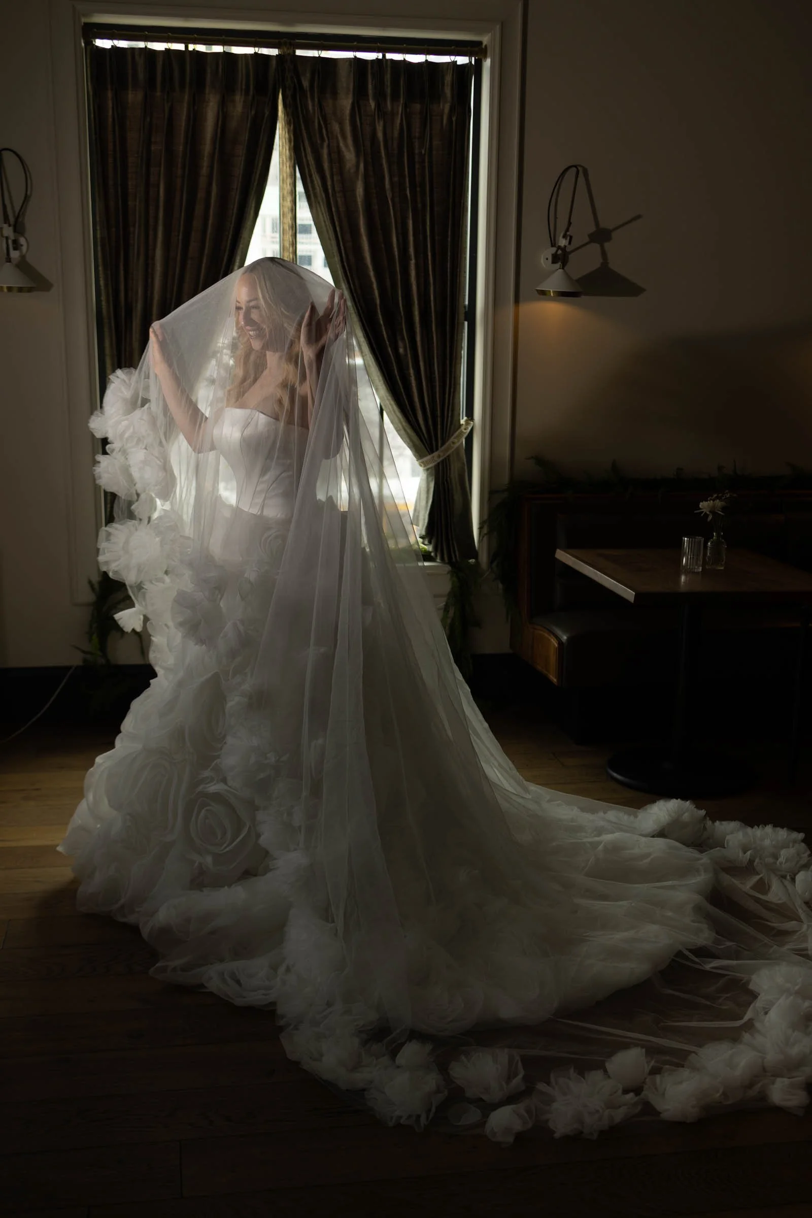  A bride in a white wedding gown with a long, flowing veil stands by a window with dark curtains, softly lit by natural light. She lifts the veil, revealing floral details on her dress. 