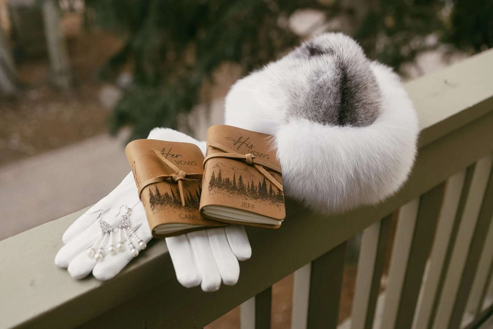  A pair of white gloves, pearl jewelry, two leather-bound vow books labeled "Her Vows" and "His Vows," and a white fur hat rest on a wooden railing, with trees blurred in the background. 