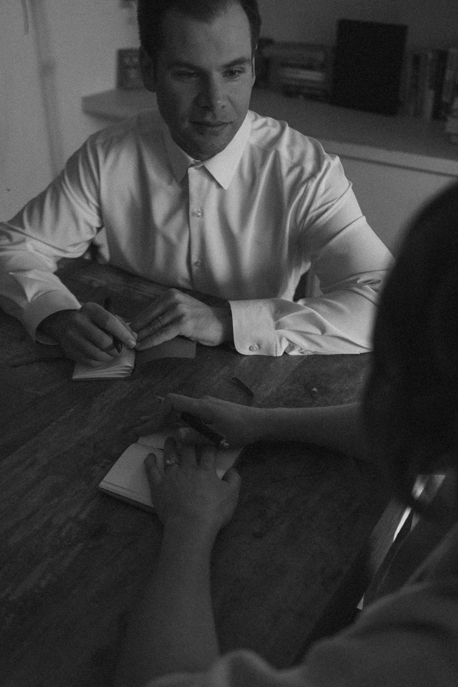  A man in a white shirt sits at a table with papers in front of him, facing another person whose hand rests on a small notebook. The scene is in black and white and appears to be a serious conversation. 