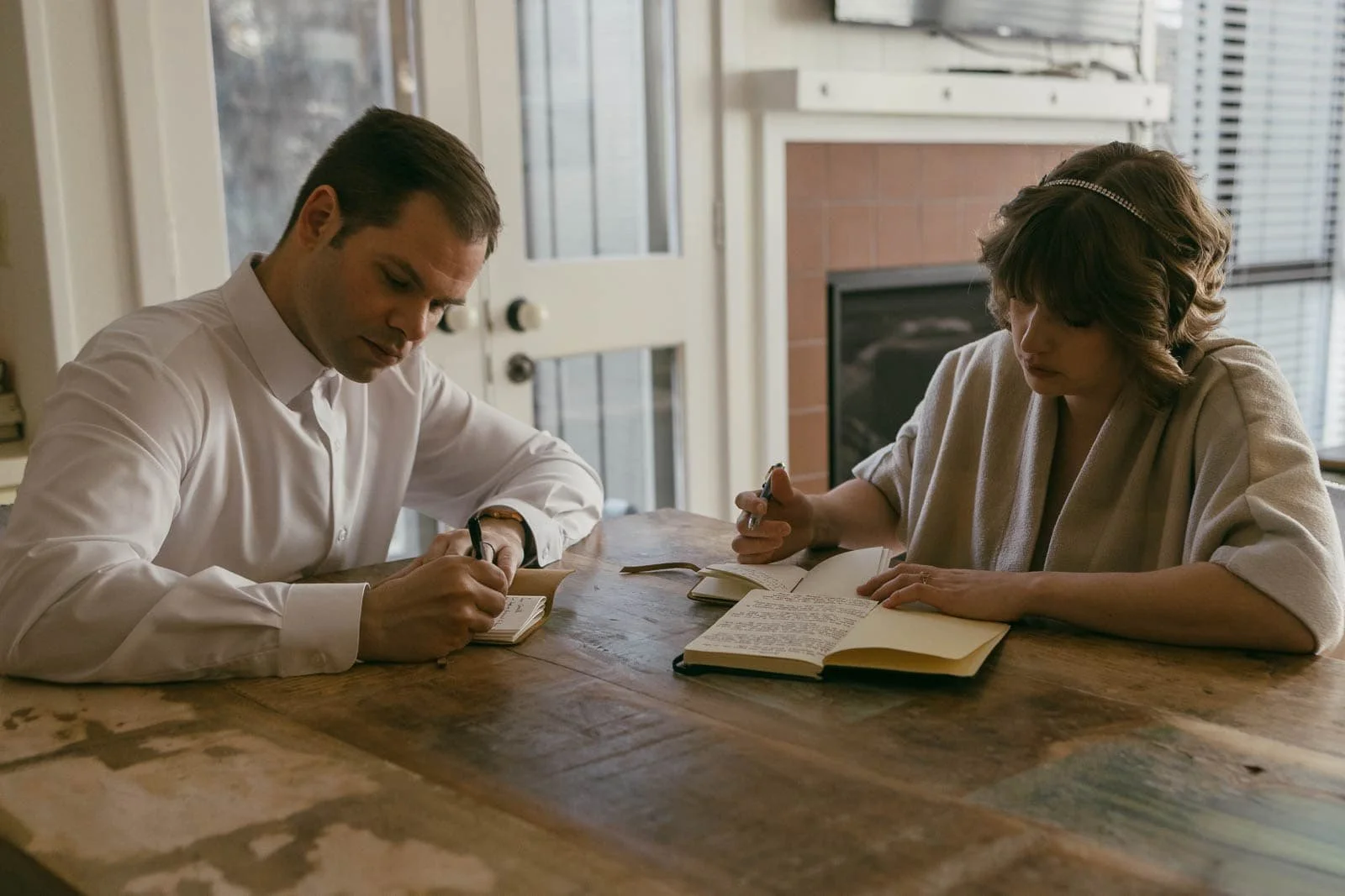 A man and woman sit together at a wooden table, both writing vows. The man wears a white shirt while the woman has short hair and a light-colored sweater. Sunlight pours through a window behind them, illuminating the room.