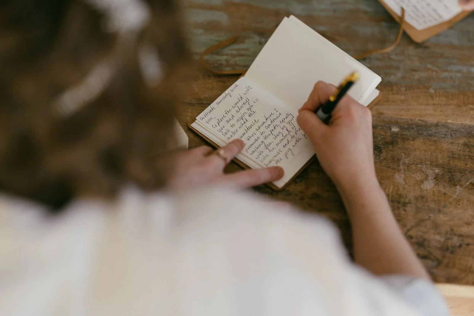  A person is writing in a small notebook with a black pen, using their left hand to hold the page open. The notebook is on a rustic wooden table, and handwritten text fills the visible page. 