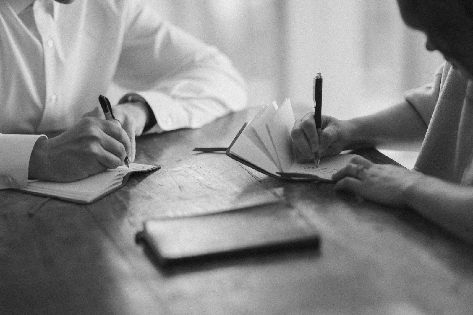  Two people sit at a wooden table, each writing in a notebook. Only their hands and arms are visible, and the photo is in black and white, creating a focused, contemplative atmosphere. 