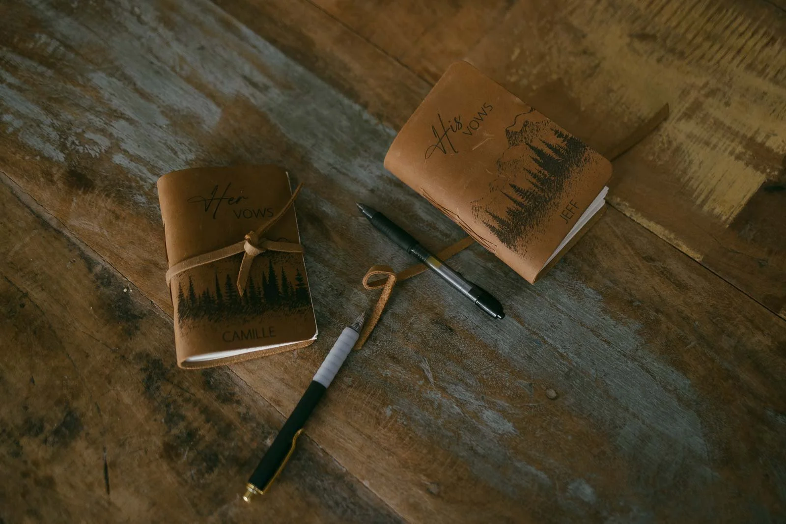  Two brown leather vow books labeled "Her Vows" and "His Vows" with names Camille and Jeff, along with two pens, placed on a rustic wooden surface. 