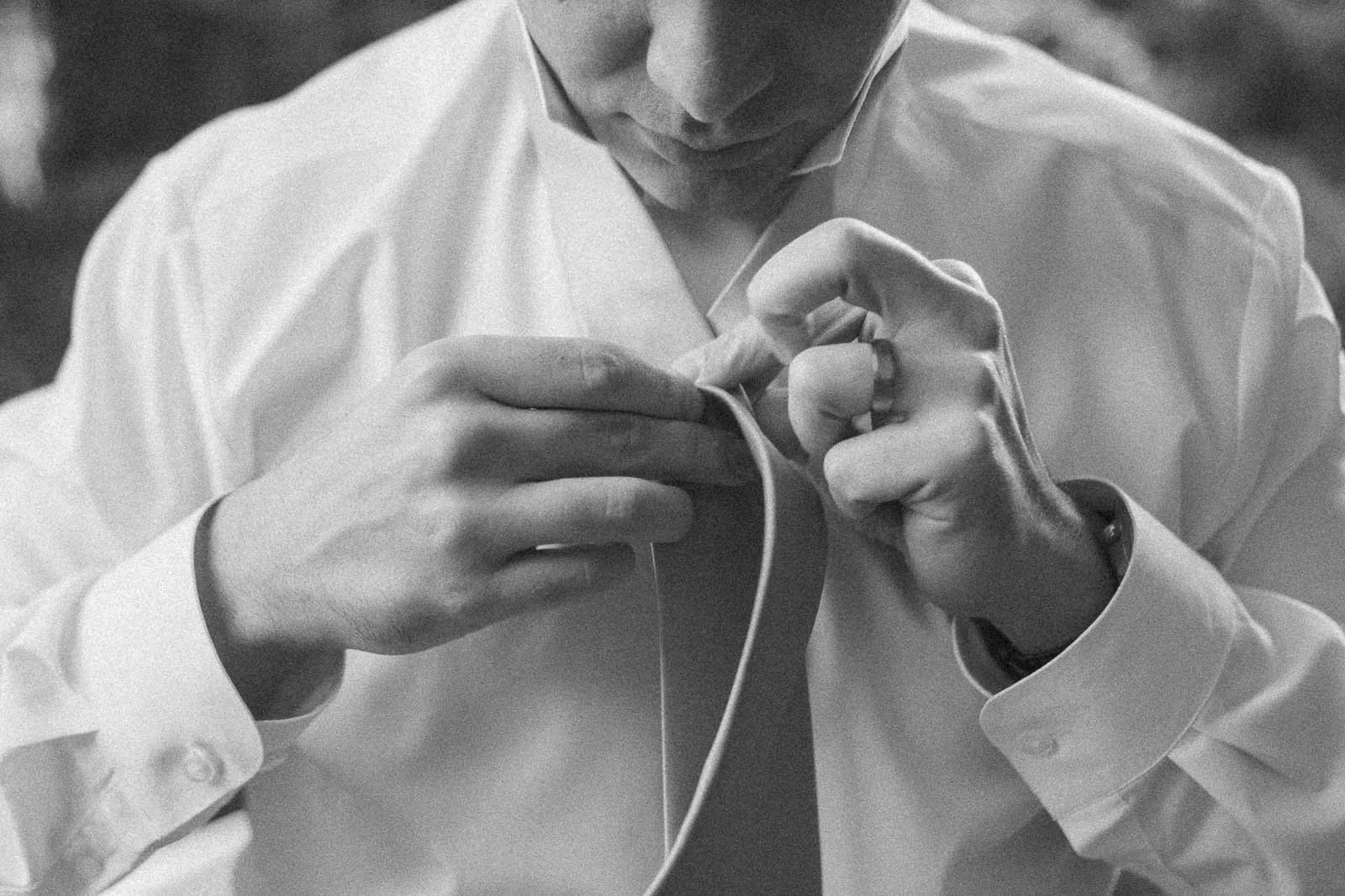  A person in a white dress shirt is tying a necktie, focusing intently on the task. The image is black and white, showing only the person’s hands, torso, and part of their face. 